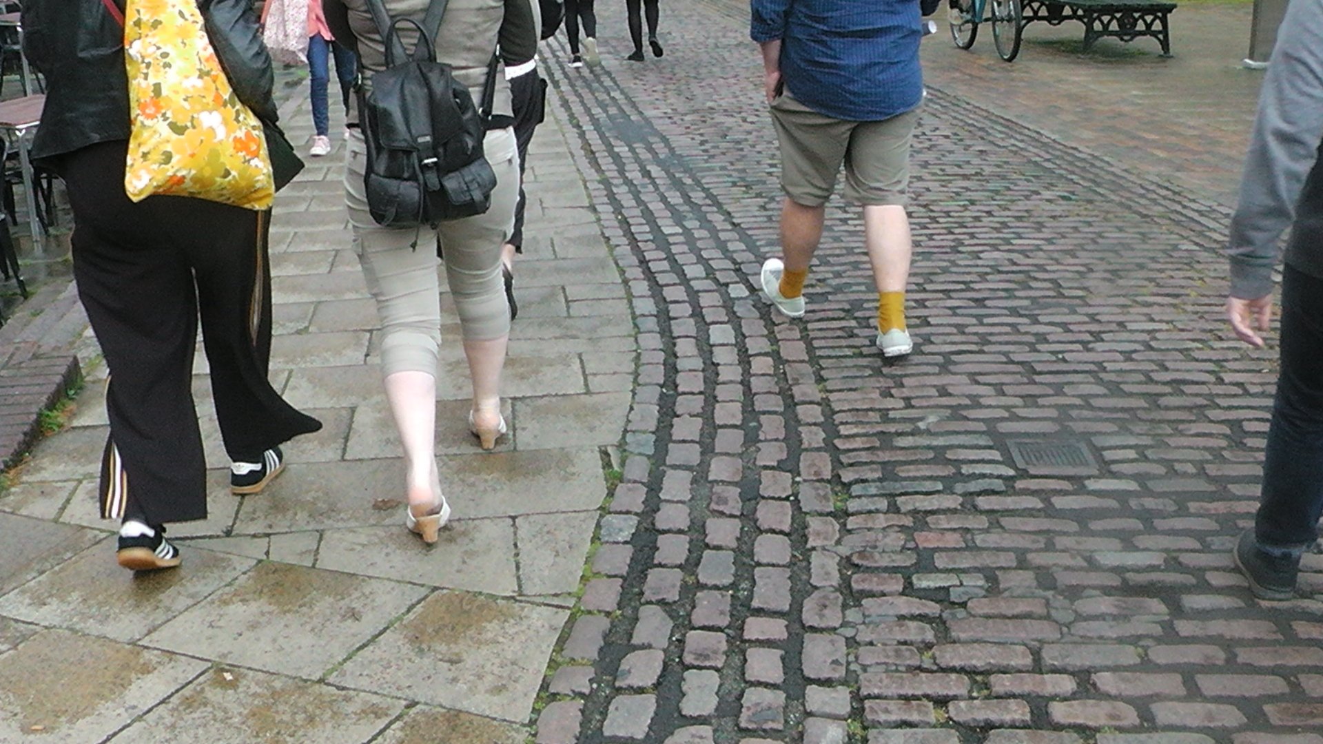 People walking on a mixed cobblestone and paved sidewalk, with benches visible in the background, in an outdoor public area.