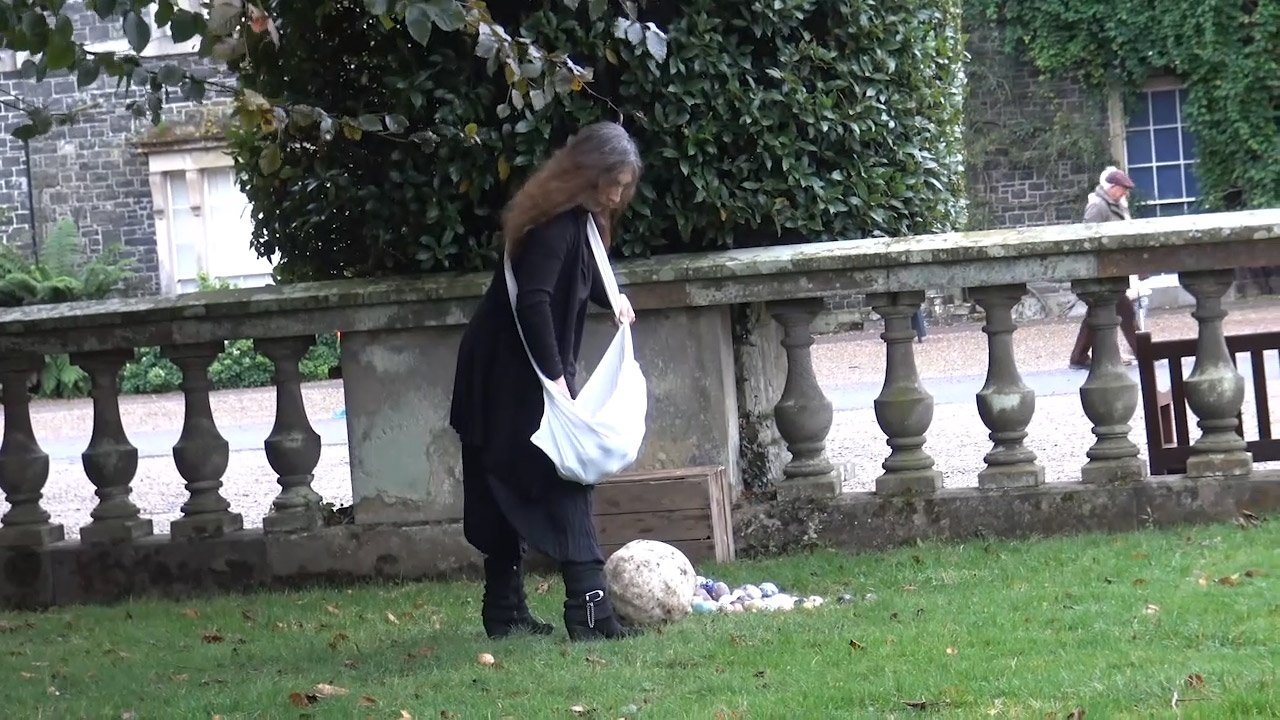 Woman in a black coat and boots placing ceramic balls around a large ceramic ball on a grassy area, with a stone fence and trees in the background.