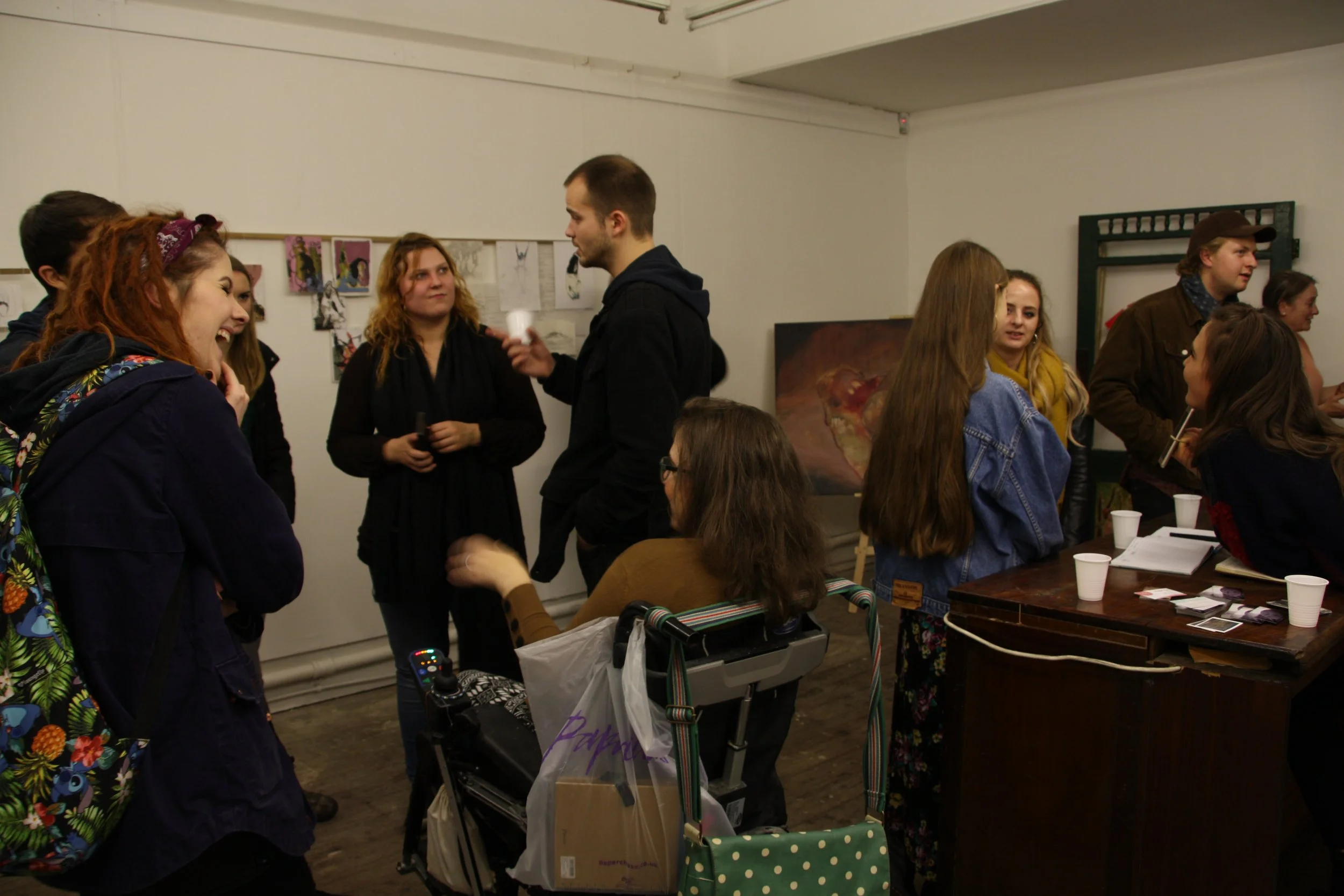 A group of people gathered indoors, some talking and smiling, with artwork hanging on the wall behind them and a table with cups and papers in the foreground.