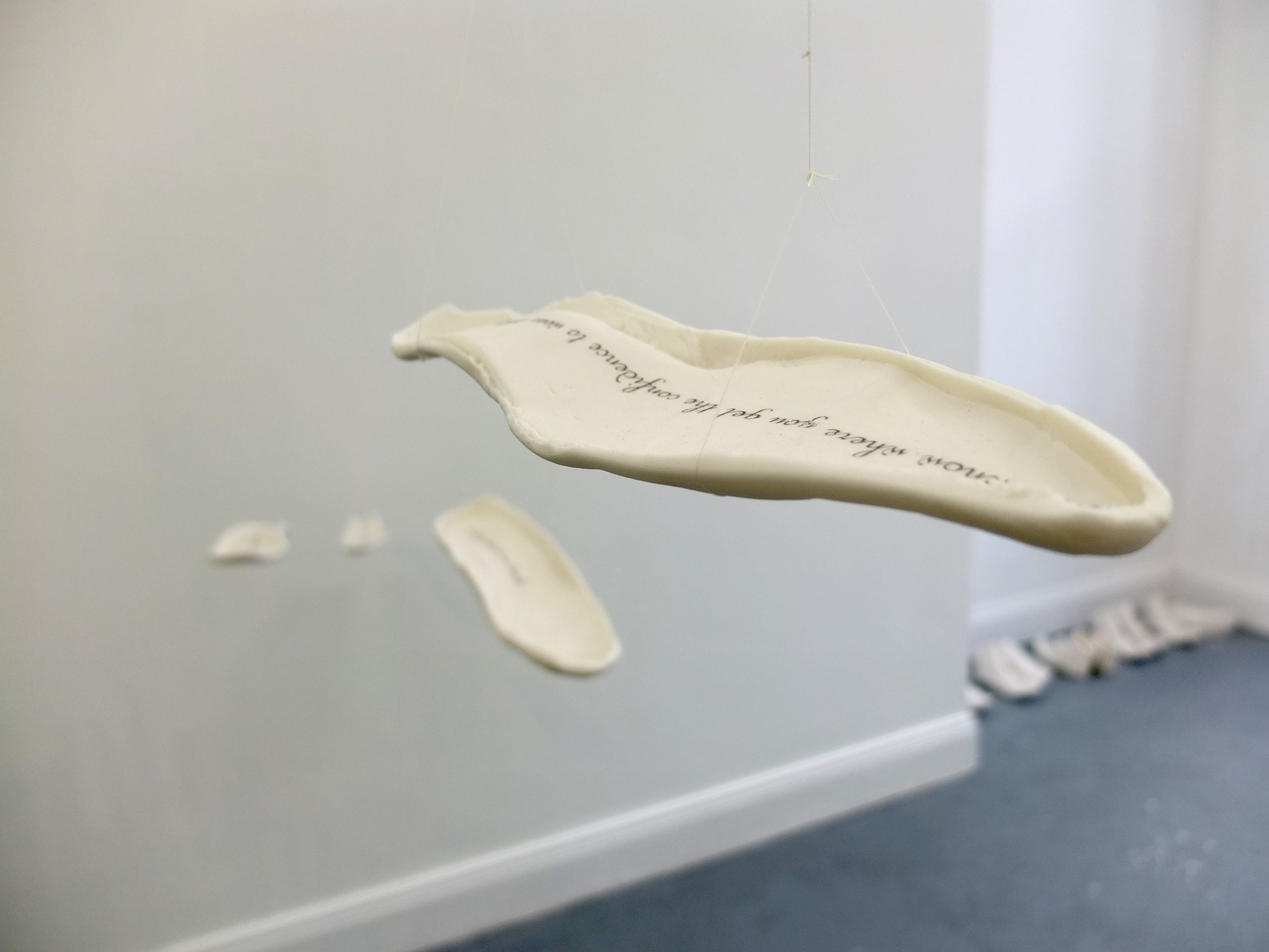 A white ceramic or porcelain , with an inscription on it that is partially visible and credits a person named Madeleine. The sculpture is mounted on a light-colored wall, and it is surrounded by smaller fragments or pieces of similar material placed along a surface below it.