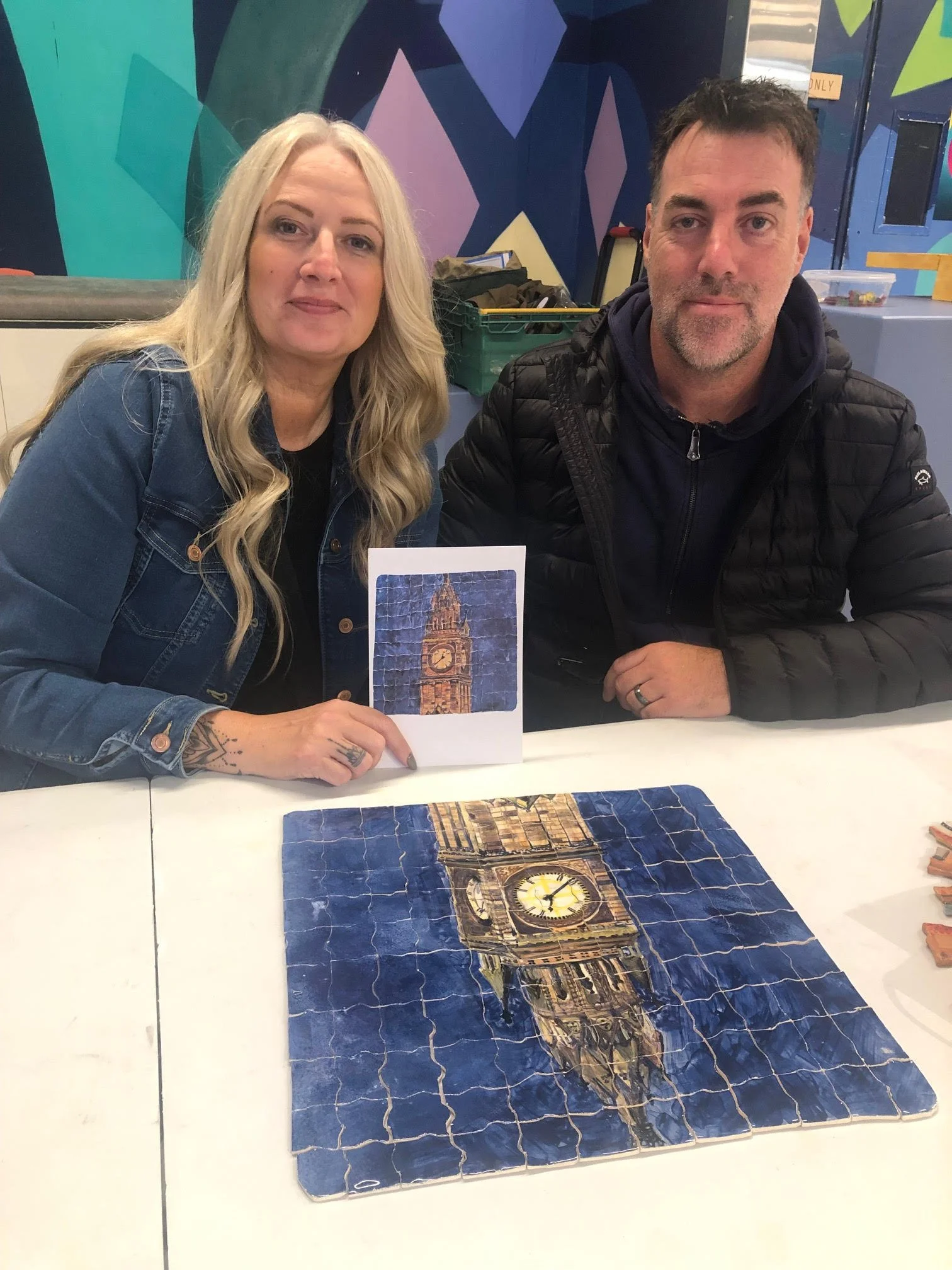 Two people sitting at a table with a large puzzle of Big Ben clock tower in progress. One person is holding a card with an image of Big Ben.
