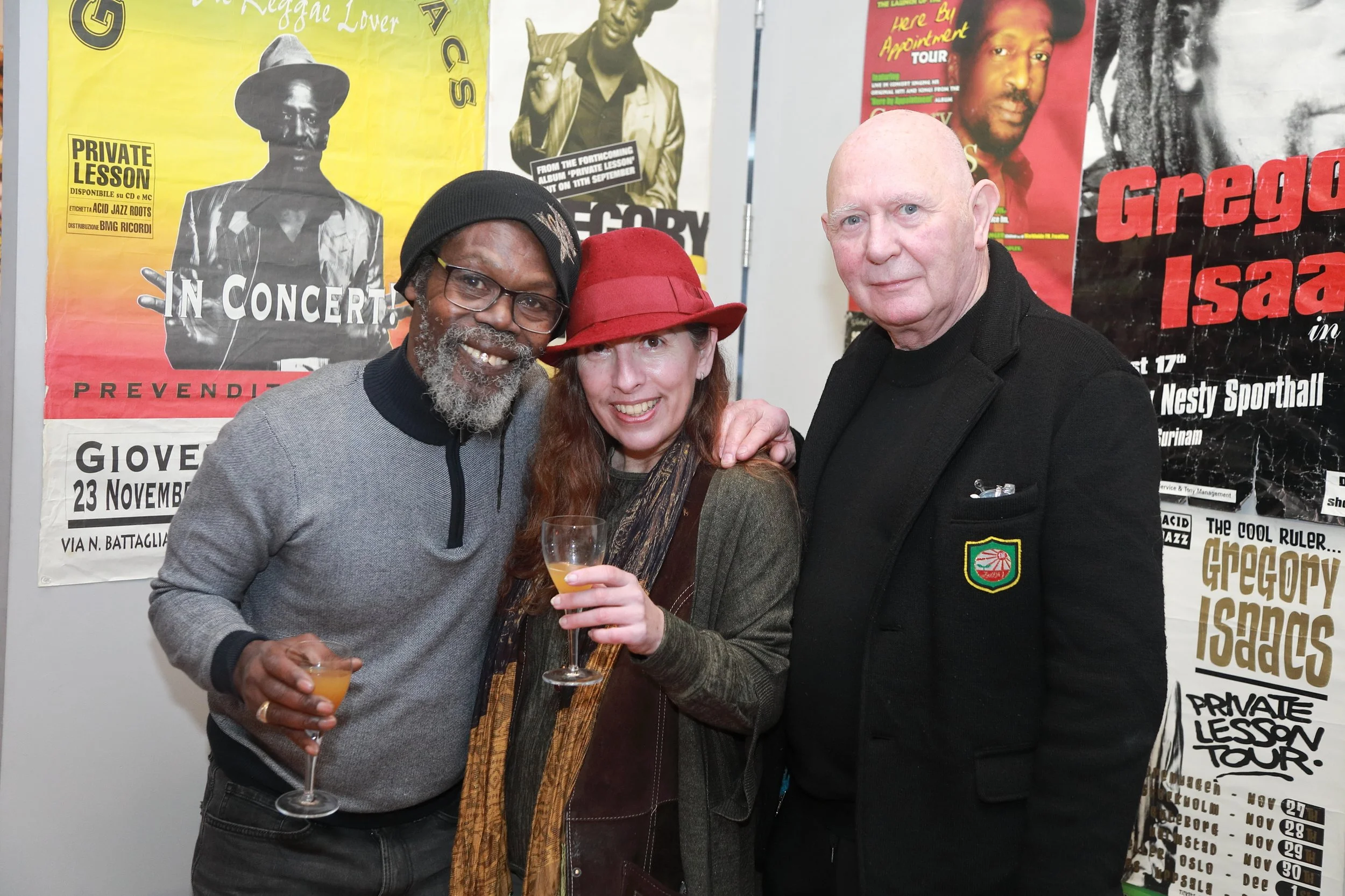 A group of three people standing together at a music event, smiling and holding drinks. The background features colorful posters advertising concerts.