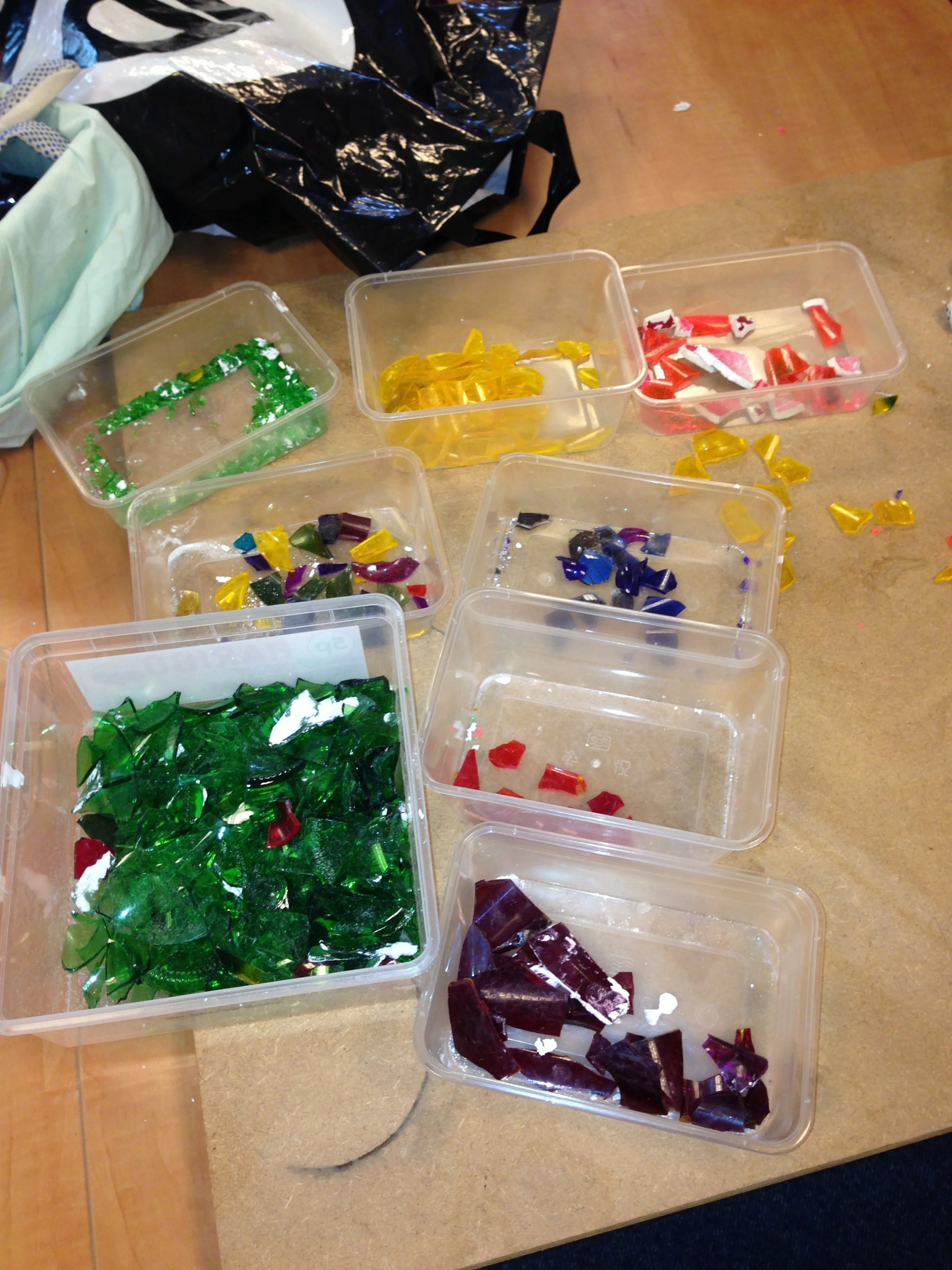 Plastic containers filled with colorful glass pieces, including green, yellow, red, purple, and dark glass shards, on a wooden table with some loose glass pieces around.