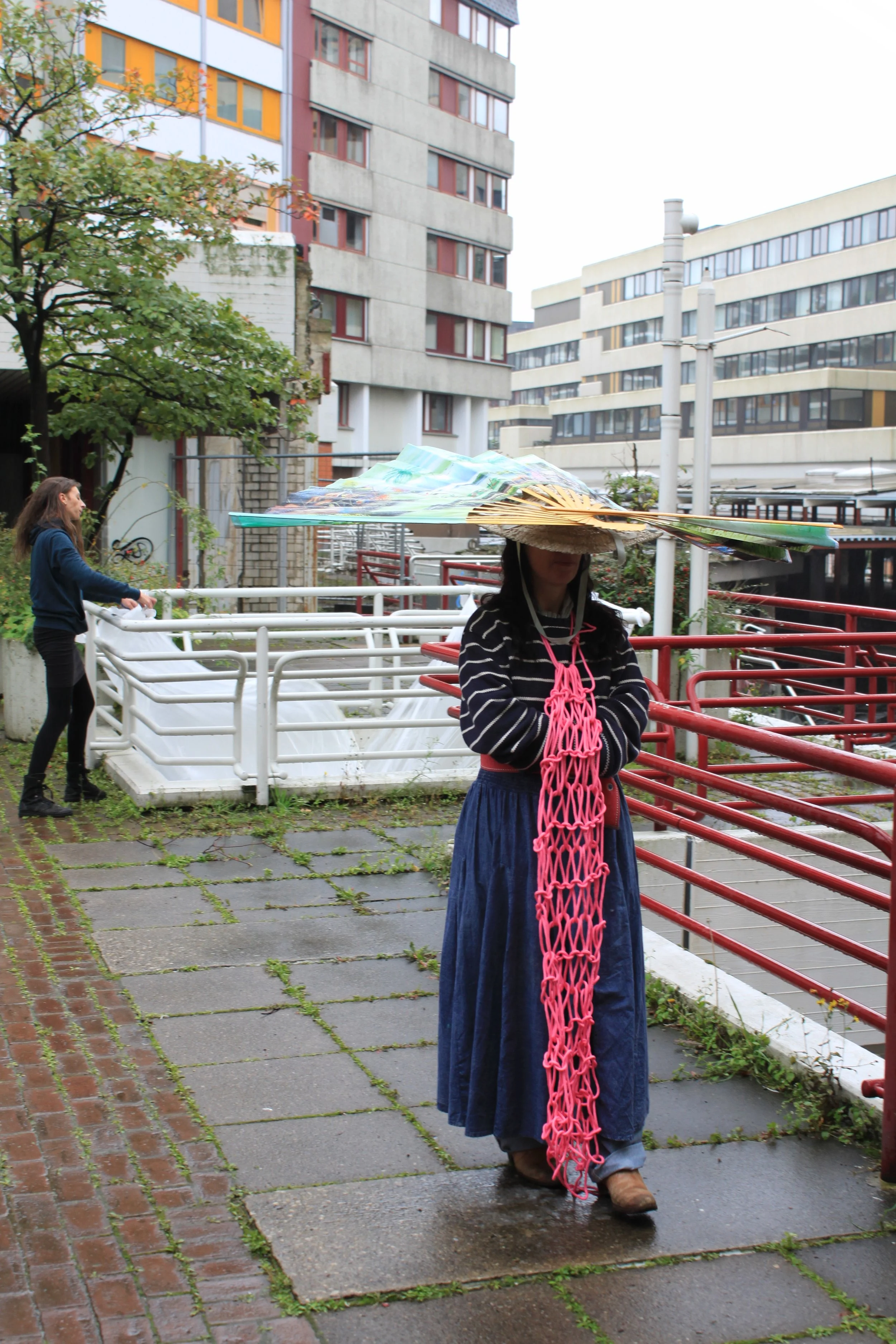 A woman wearing a vintage dress and a large, decorative hat stands on a wet sidewalk with red railing in an urban area. There are buildings with multiple windows, trees, and another woman walking in the background.