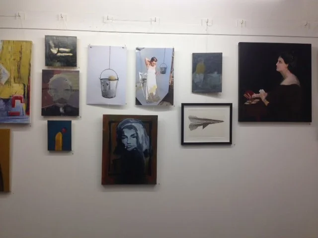 Gallery wall with various paintings including abstract art, a woman in a white dress, a portrait of a woman, a boat, and a person holding a red object.
