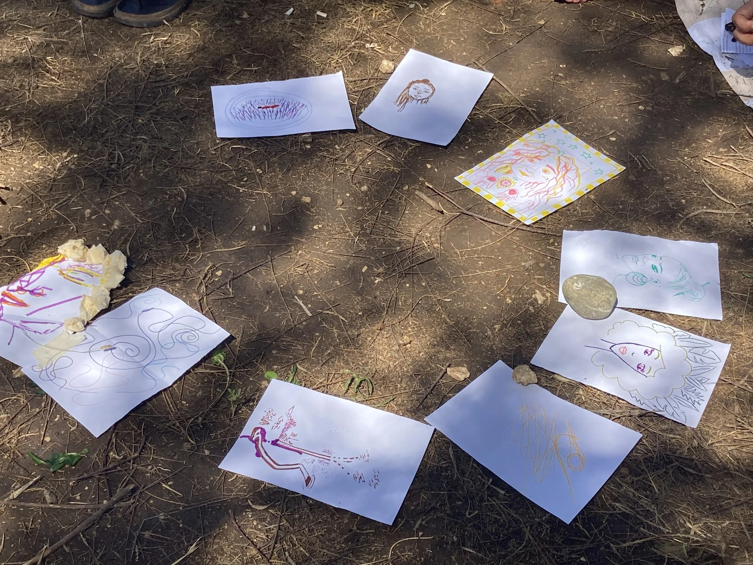 Colorful children's drawings displayed on the ground outdoors, some taped to rocks, featuring various characters and abstract designs.