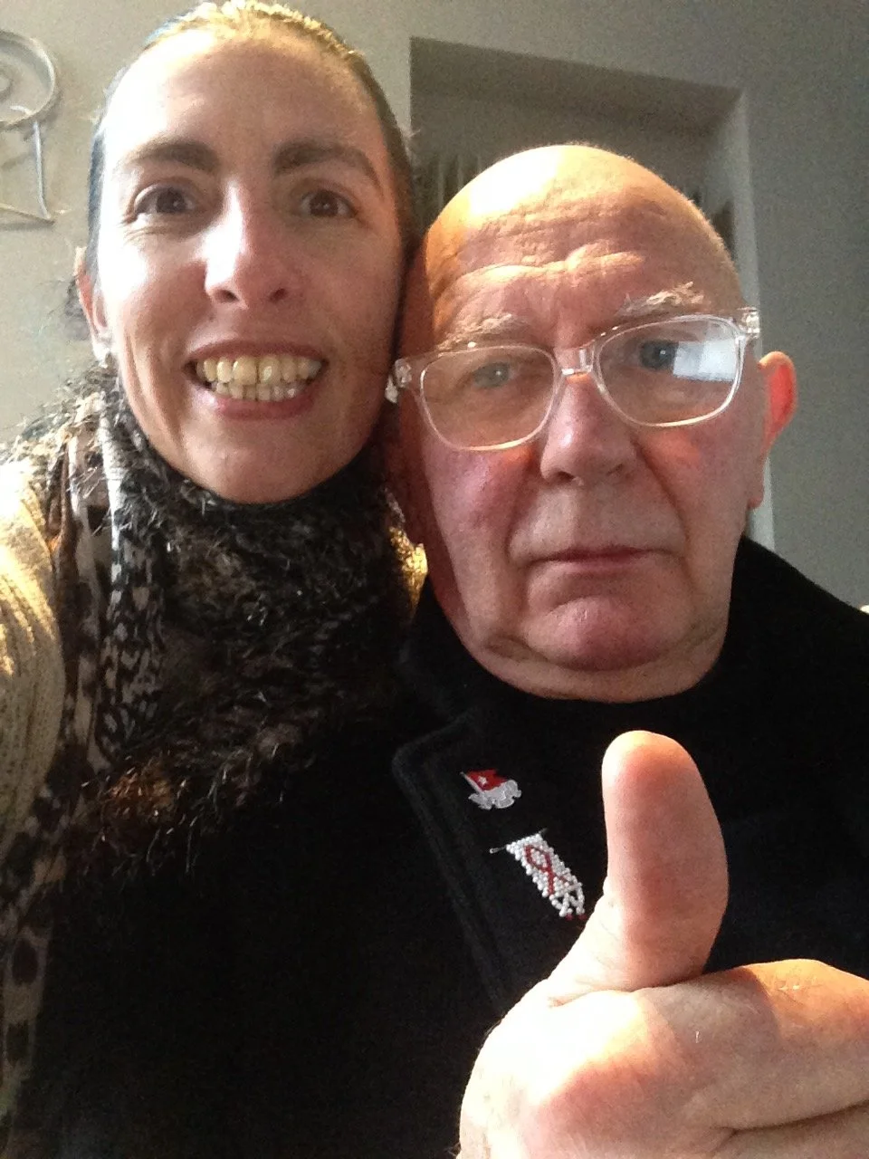 A woman and an older man taking a close-up selfie indoors. The woman is smiling and sticking her tongue out, with her arm around the man's shoulder. The man is wearing glasses and giving a thumbs-up. They appear to be in a cozy setting with a staircase in the background.
