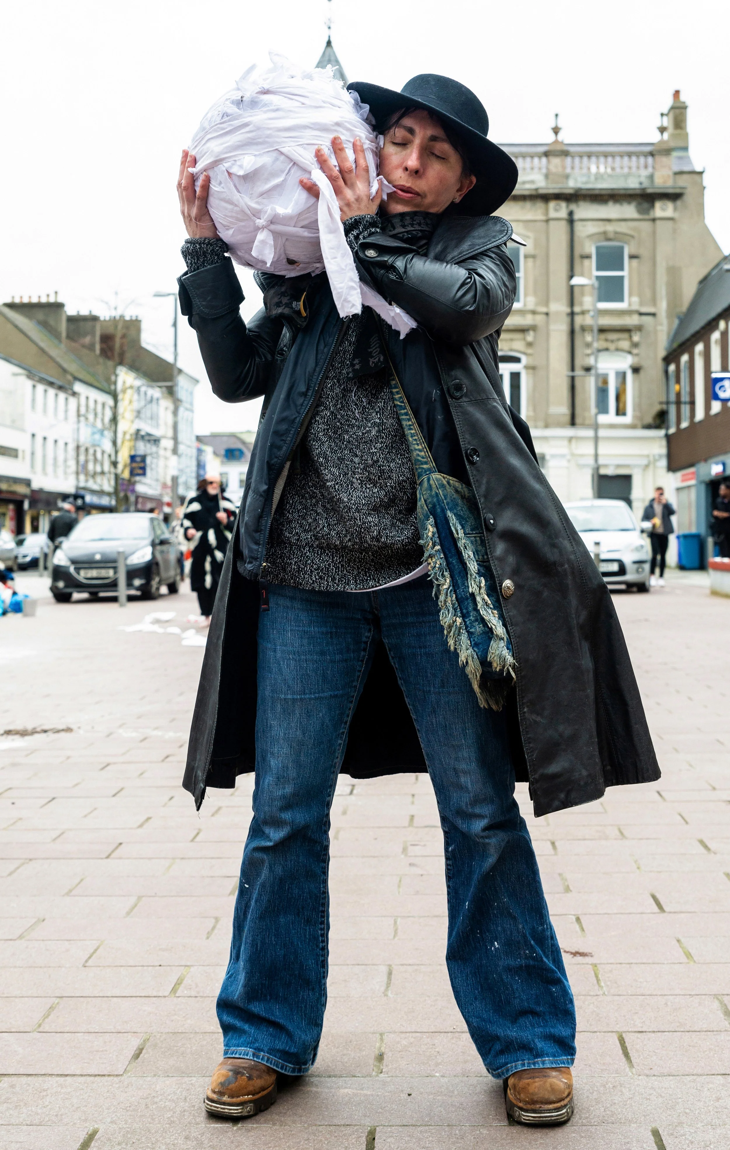 A person wearing a black hat, black leather jacket, and blue jeans is standing on a street holding a large bundle wrapped in white cloth. The person is embracing the bundle with their eyes closed. The background shows buildings, people walking, and p