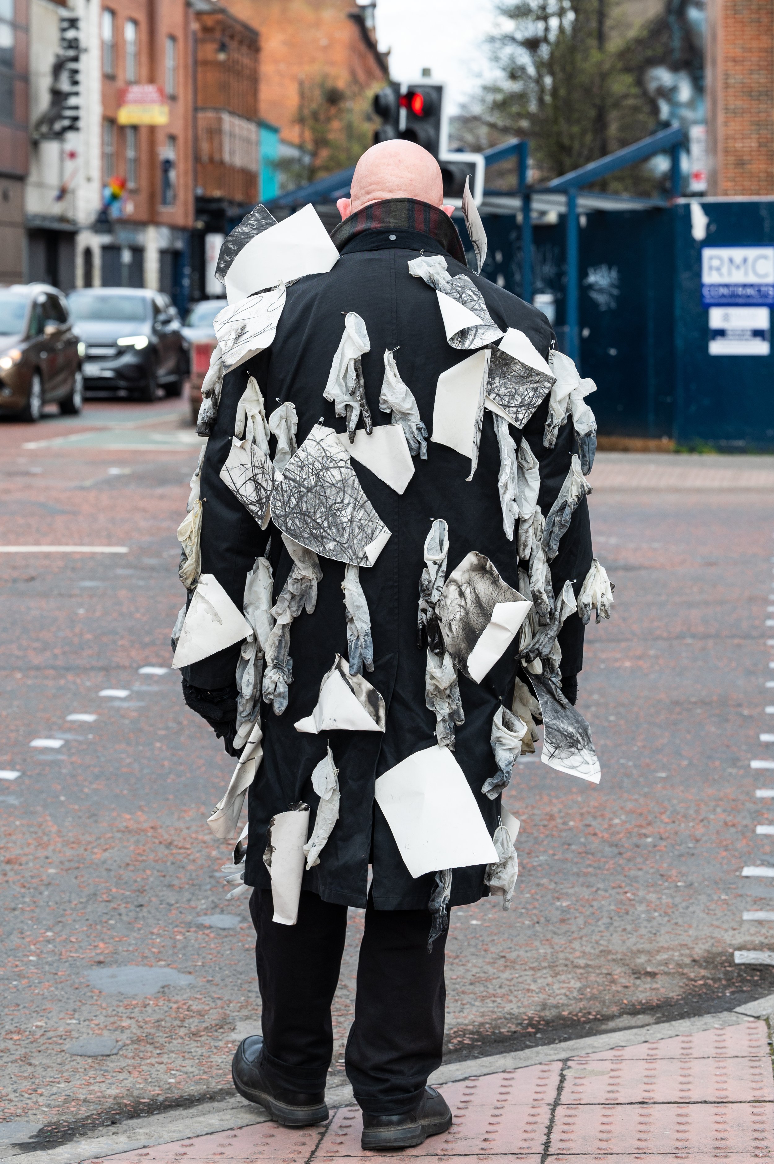 A person with a bald head facing away, wearing a black coat covered with torn pieces of paper with sketches and lines, walking across a city street with parked cars and brick buildings, red traffic light visible.