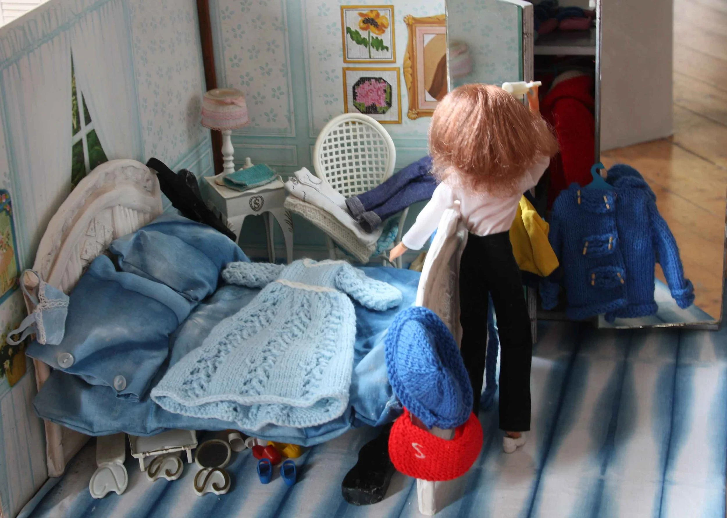 A dollhouse bedroom with a bed, a blue knitted dress draped on it and a doll standing in the room