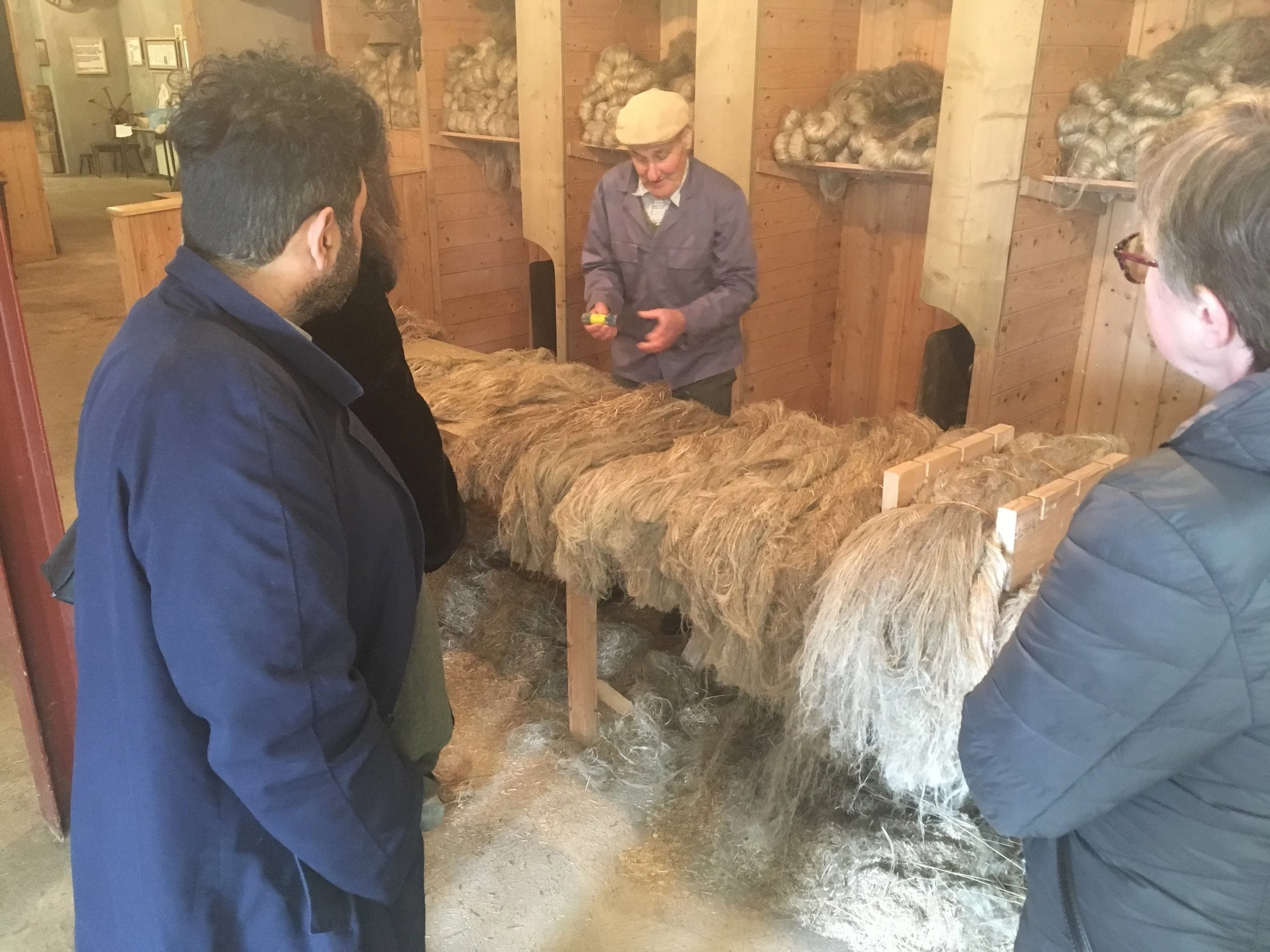 A woman explains wool processing to a group of people in a rustic wooden room, with shelves of wool in the background.