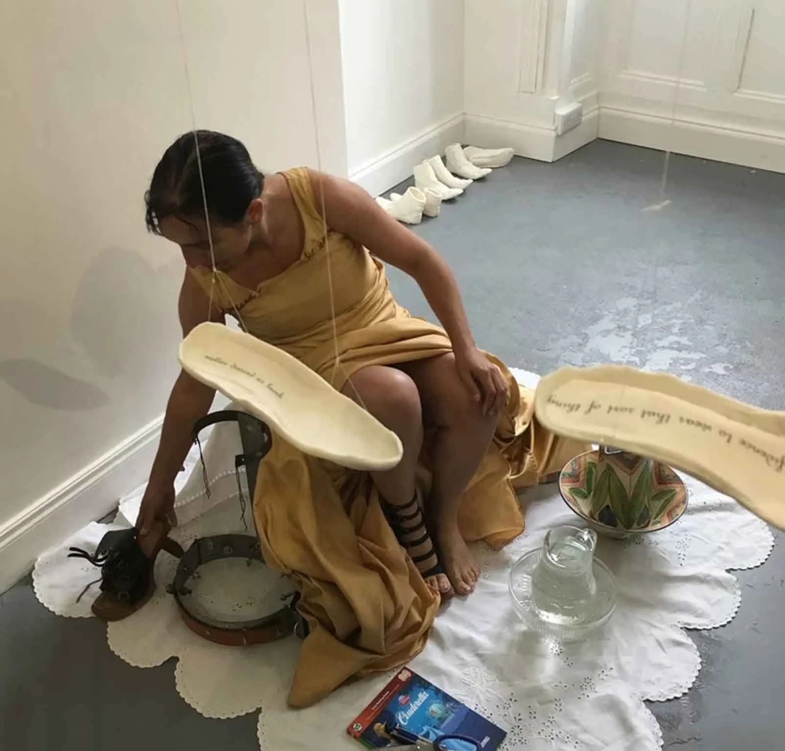 A woman dressed in a yellow gown, sitting on a white lace cloth, windows, and decorative items, with several white shoes and a water container on the floor.