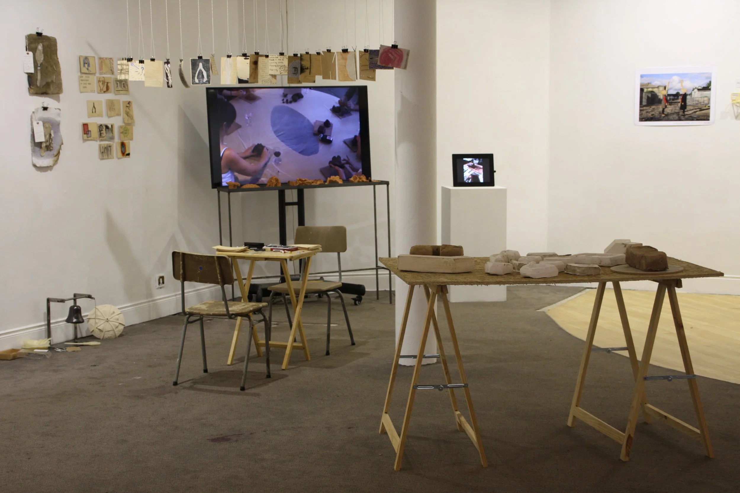 An art gallery or exhibition space with tables displaying various objects and photographs on the walls. There are chairs, a large television screen, and art pieces including hanging papers, on the wall and tables.