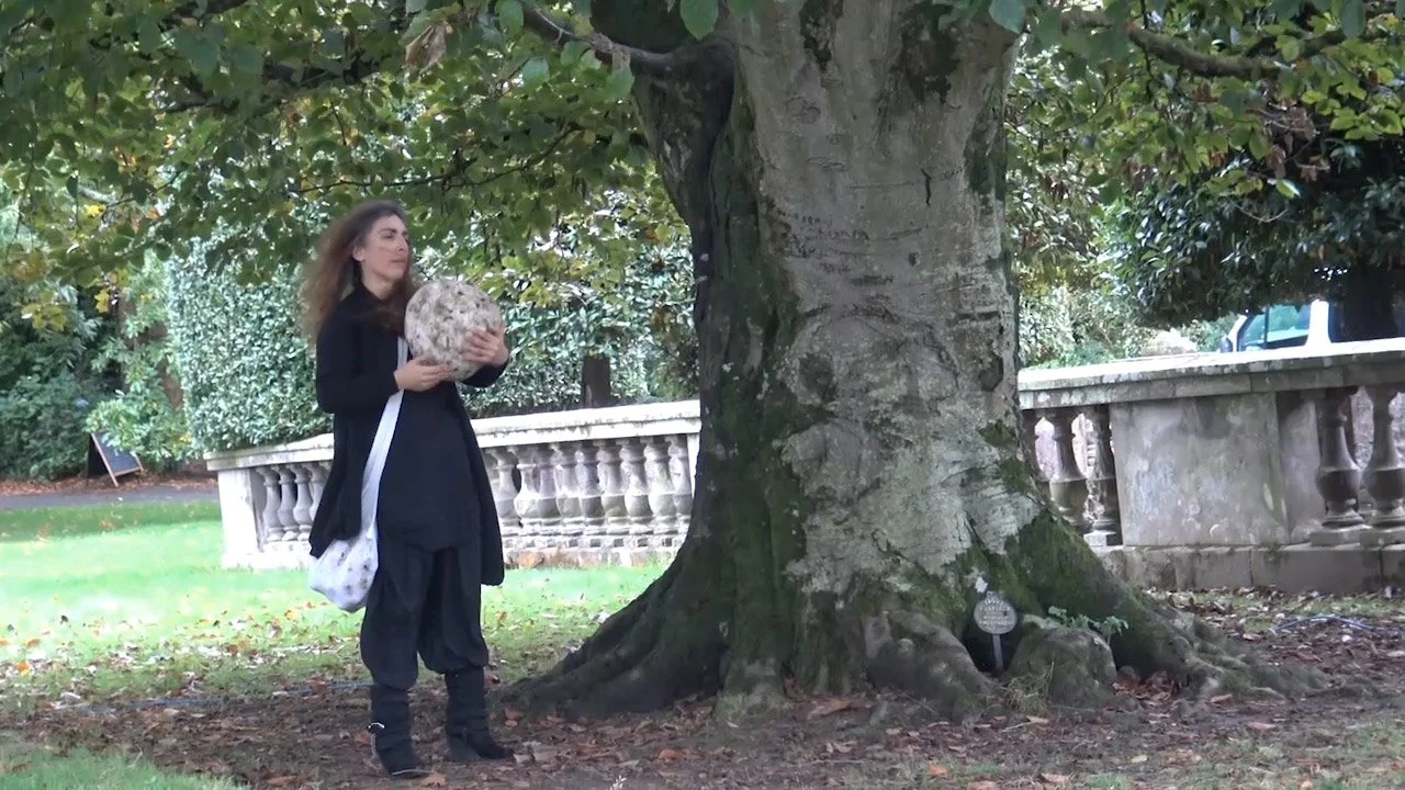 A woman holding a large, round, textured object, standing next to a large tree with a stone balustrade in the background in a park setting.