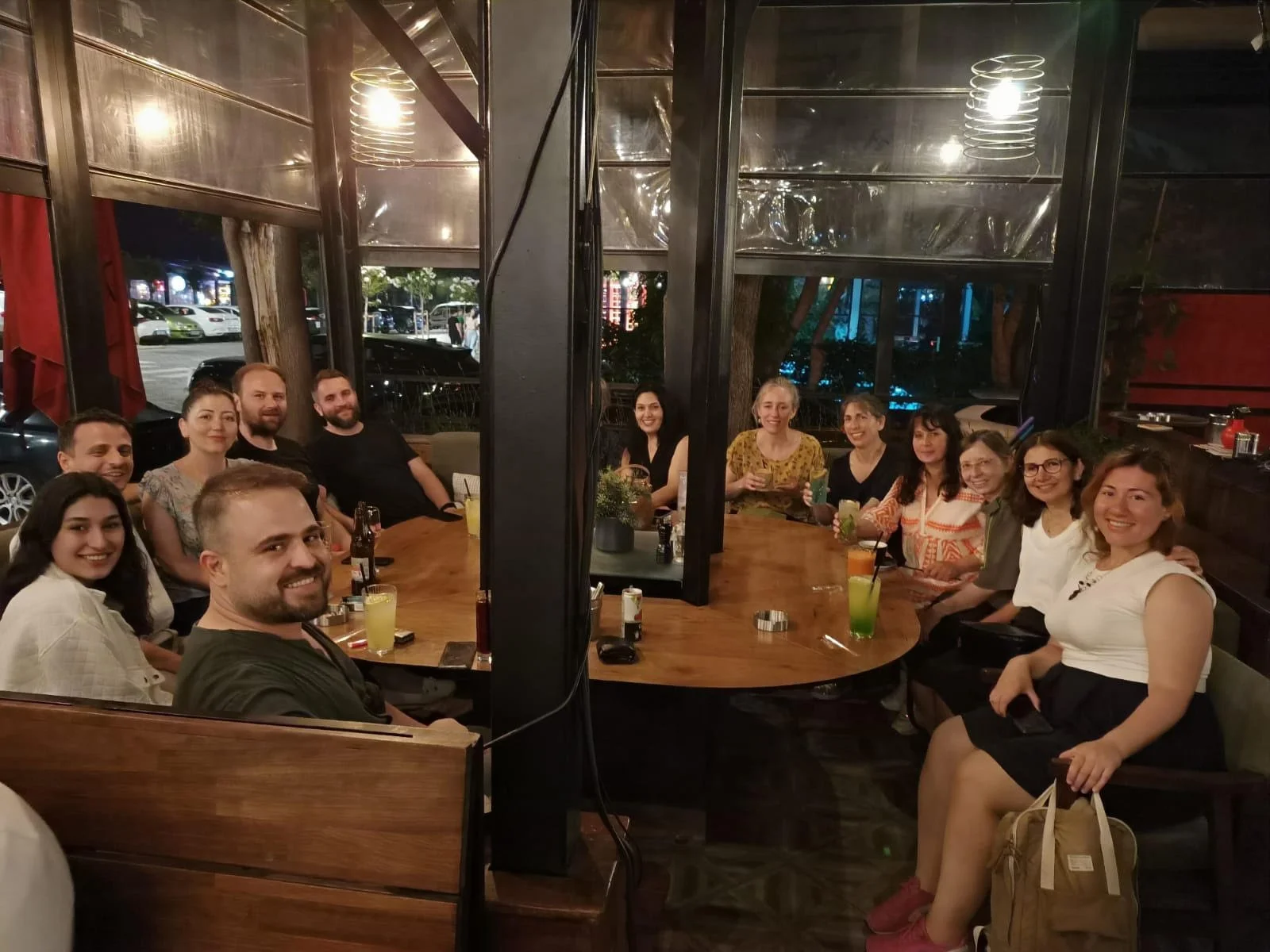 A group of twelve people sitting around a large wooden table at a restaurant or bar, smiling at the camera, with drinks in front of them, in a dimly lit indoor setting.