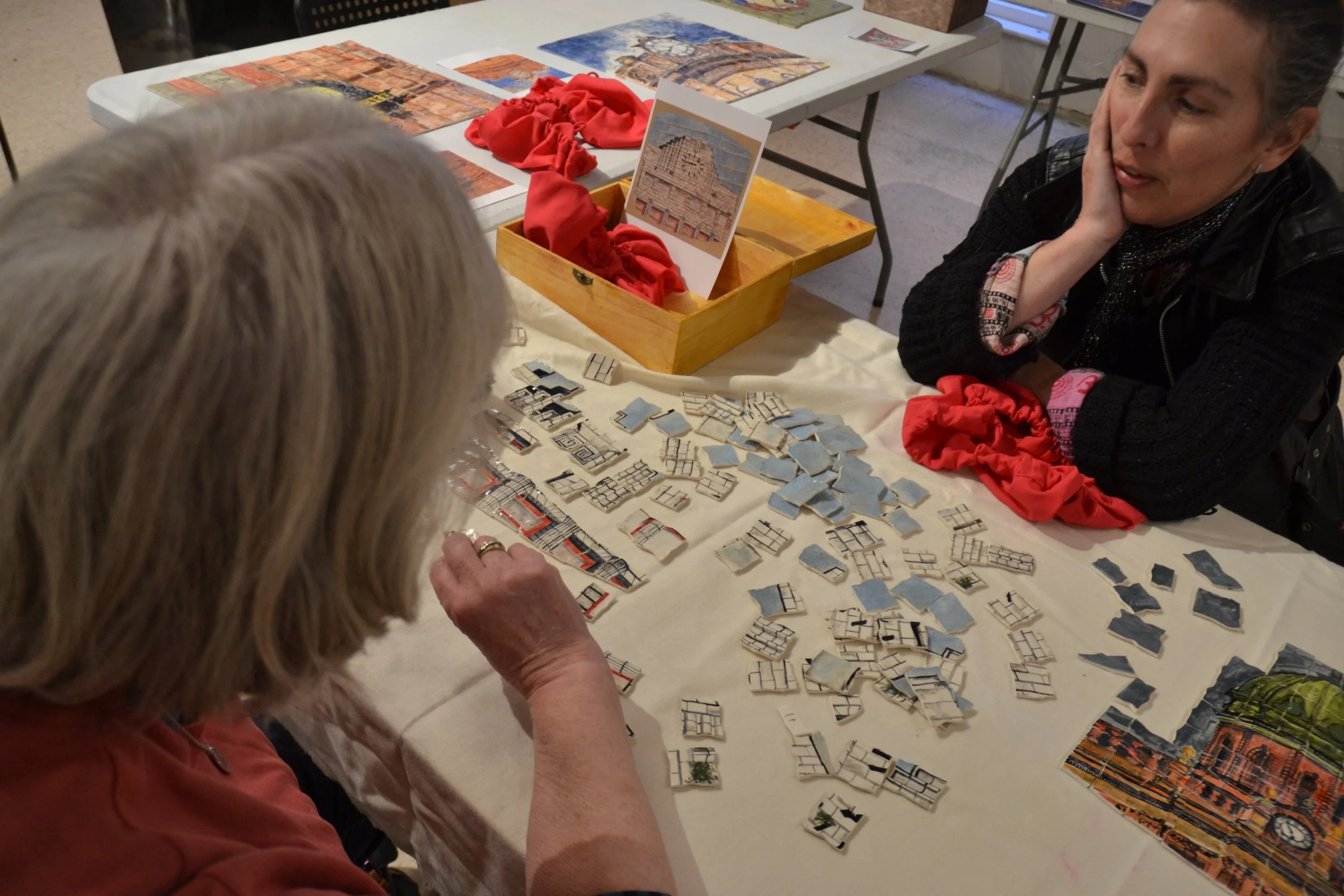 Two women are playing a tile-matching game at a table, with several cityscape puzzle pieces spread out. One woman with grey hair wearing a red shirt observes, while another woman with dark hair and a black sweater talks nearby. There are art prints a