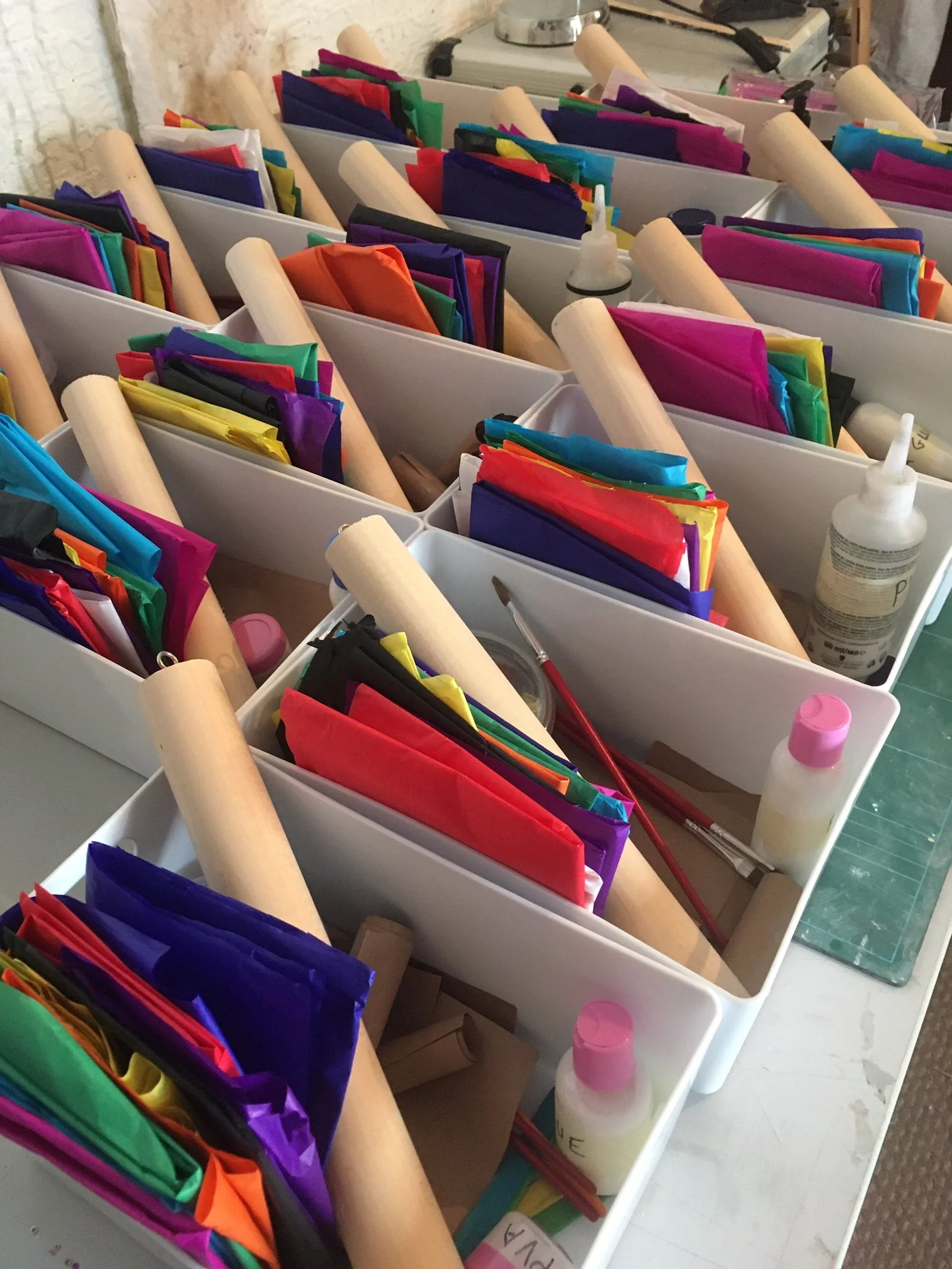 Multiple white storage bins filled with colorful folded papers, some with wooden dowels, bottles of glue, and various craft supplies on a workspace.