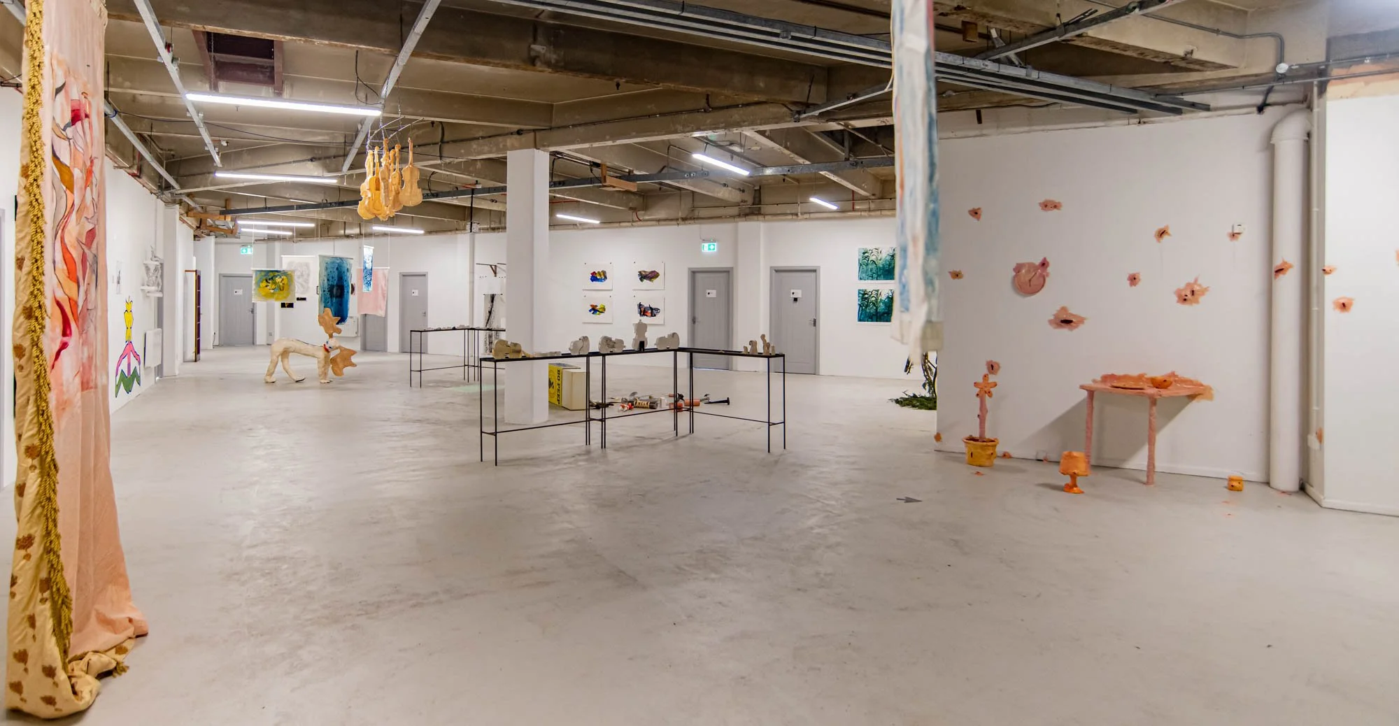Interior of an art gallery with various colorful sculptures, paintings, and artworks displayed on white walls and on black display tables, in an industrial-style space with visible ceiling pipes and concrete floors.