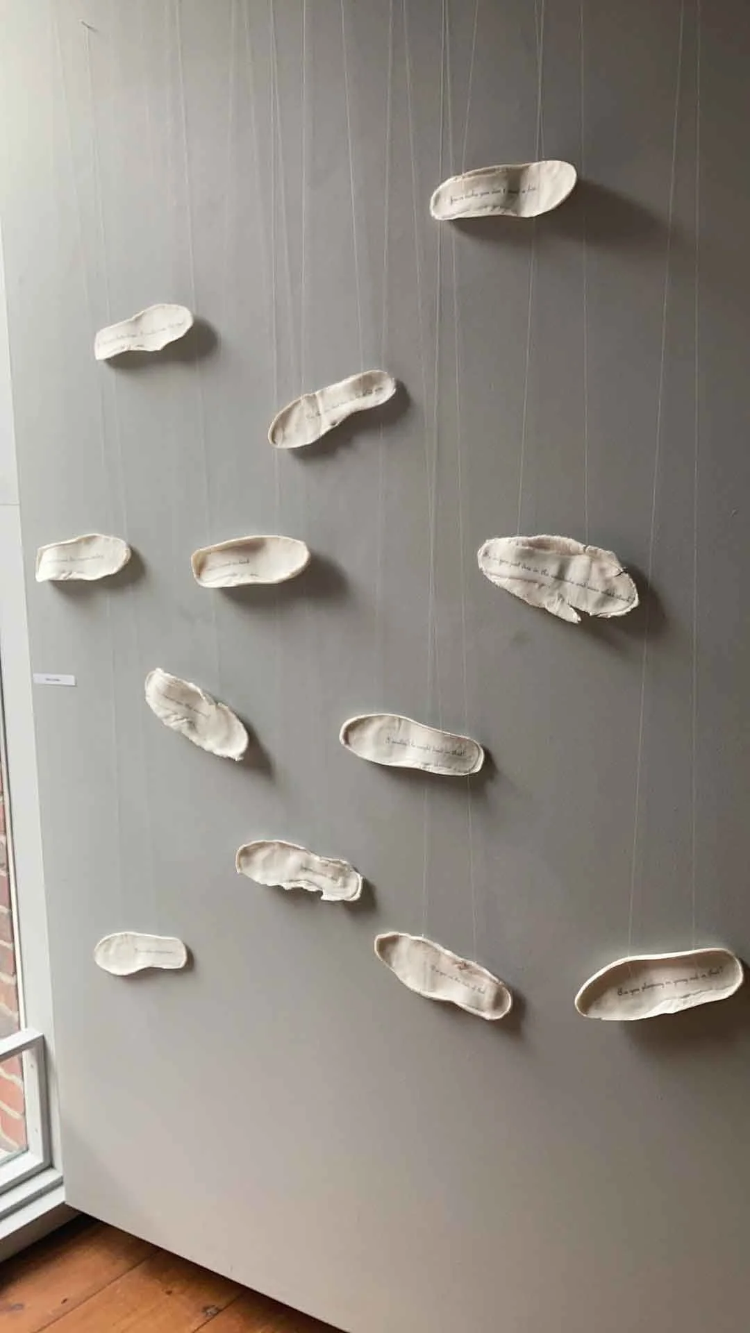 Display of multiple white ceramic footprints with handwritten text, hung by strings on a gray wall.