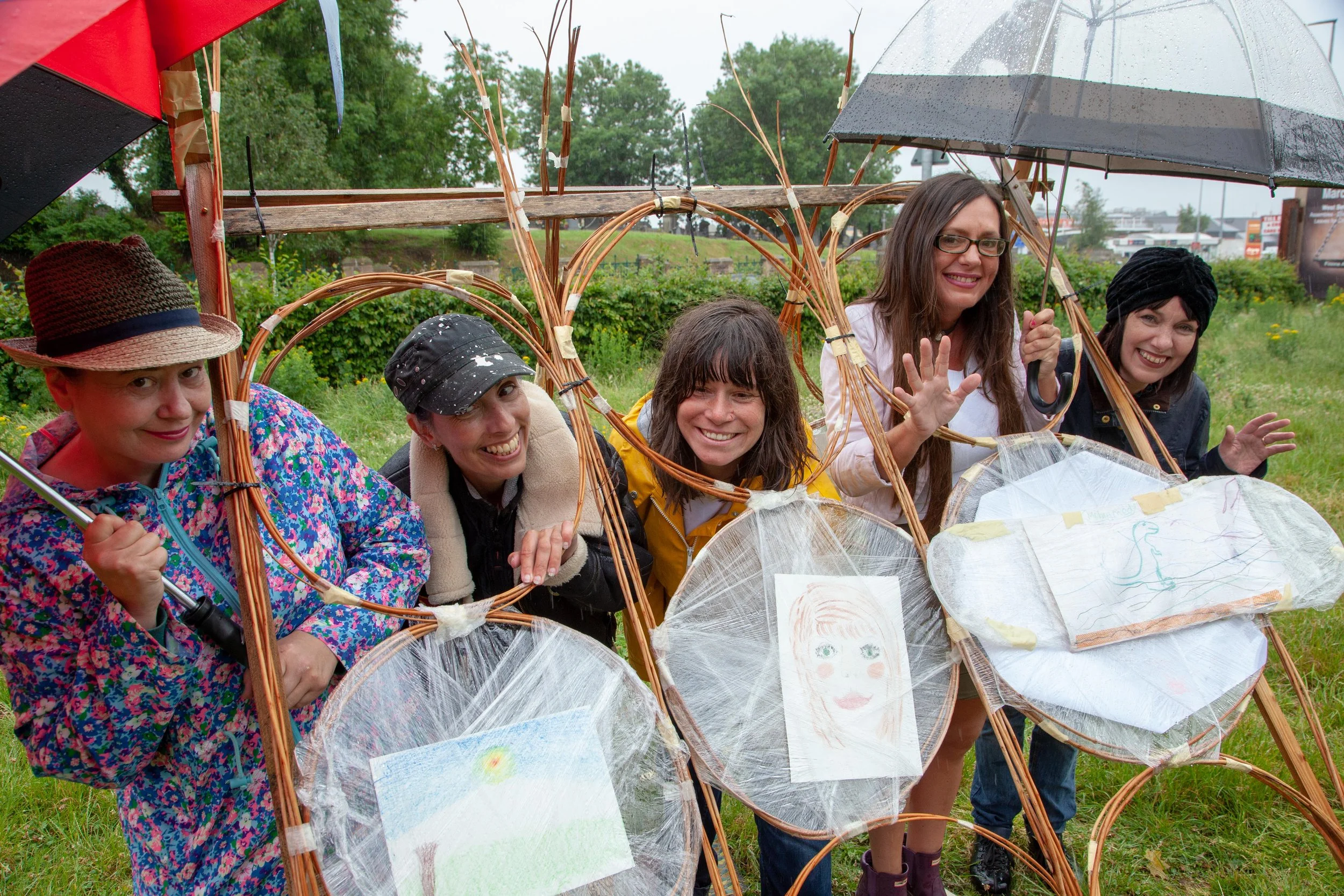 Group of women outdoors in rainy weather, smiling and holding umbrellas, standing behind a display of framed children's artwork on bamboo and string structures.