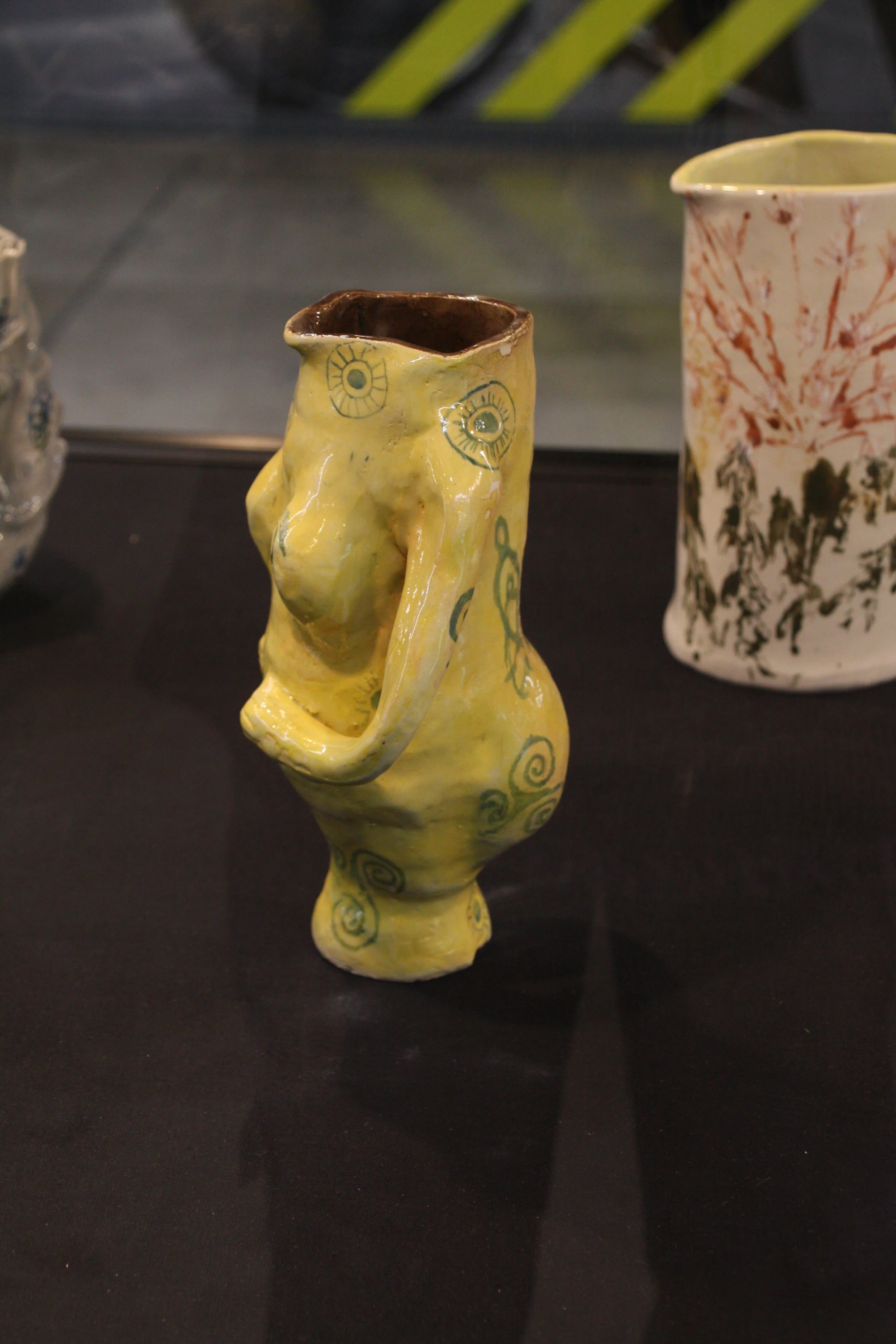 A yellow ceramic vase with a face-like design featuring eye details and spiral patterns, with a second taller vase decorated with tree branches in the background.