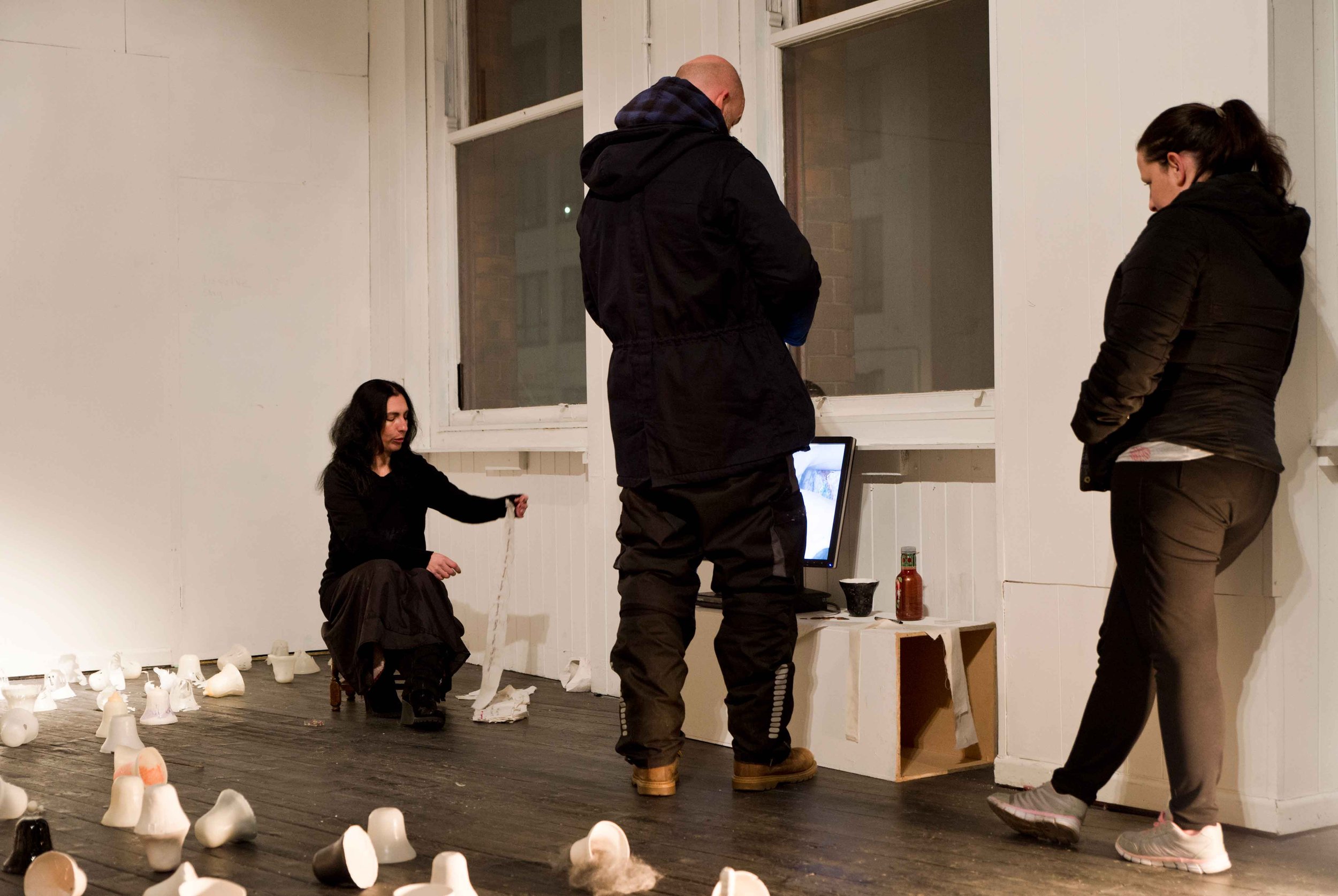 People in black jackets standing and sitting around a laptop in a room with white walls and large windows, surrounded by scattered ceramic objects on the floor.