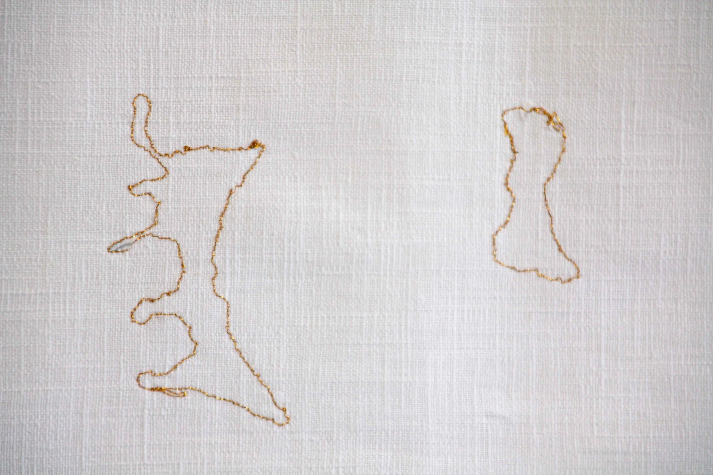 Close-up photo of a white fabric with a gold thread embroidery outlining the shape of the state of Alaska.
