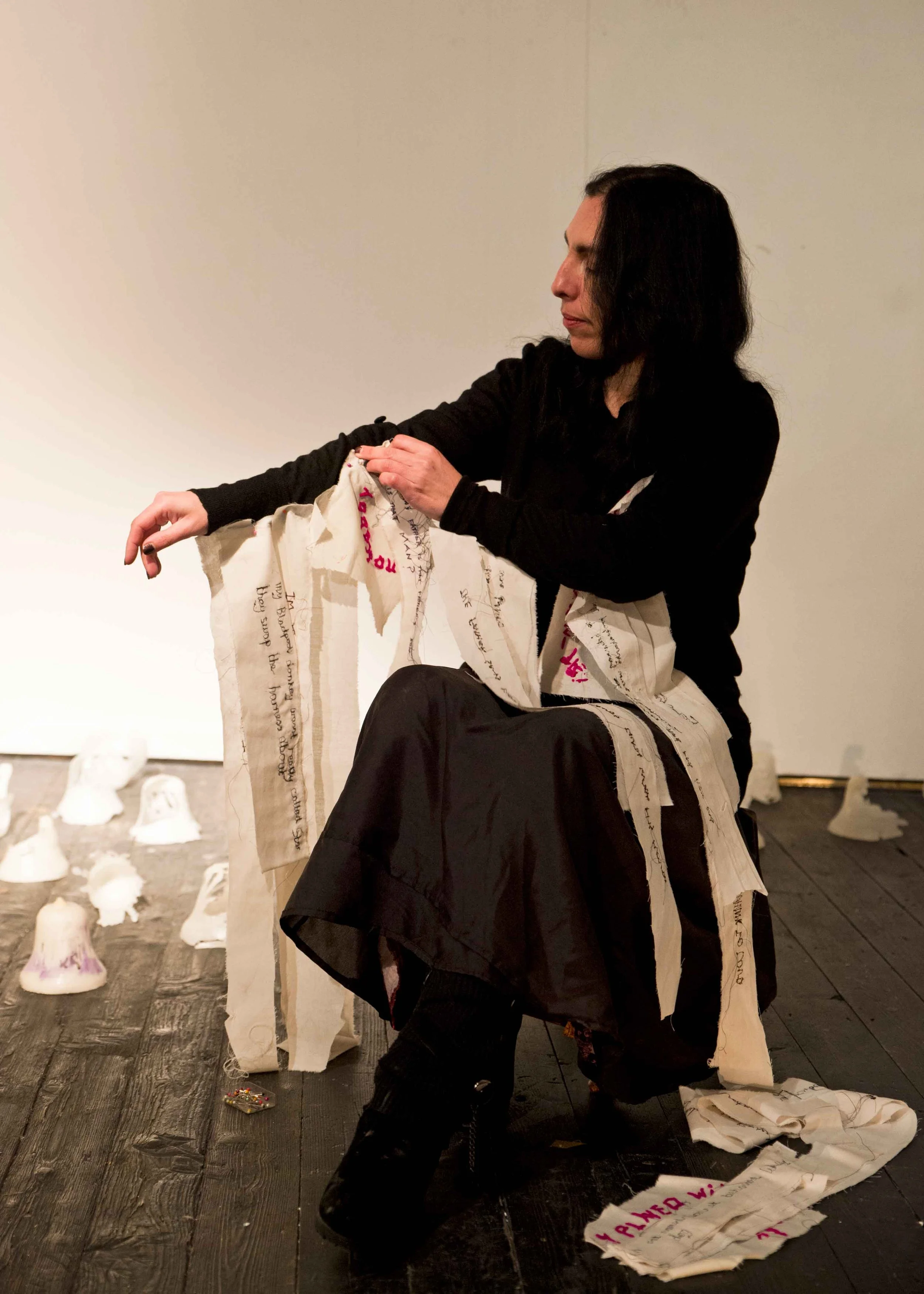 A woman with black hair wearing black clothing sitting on a wooden floor, holding a torn piece of fabric with handwritten text, with scattered fabric pieces and white ceramic objects in the background.