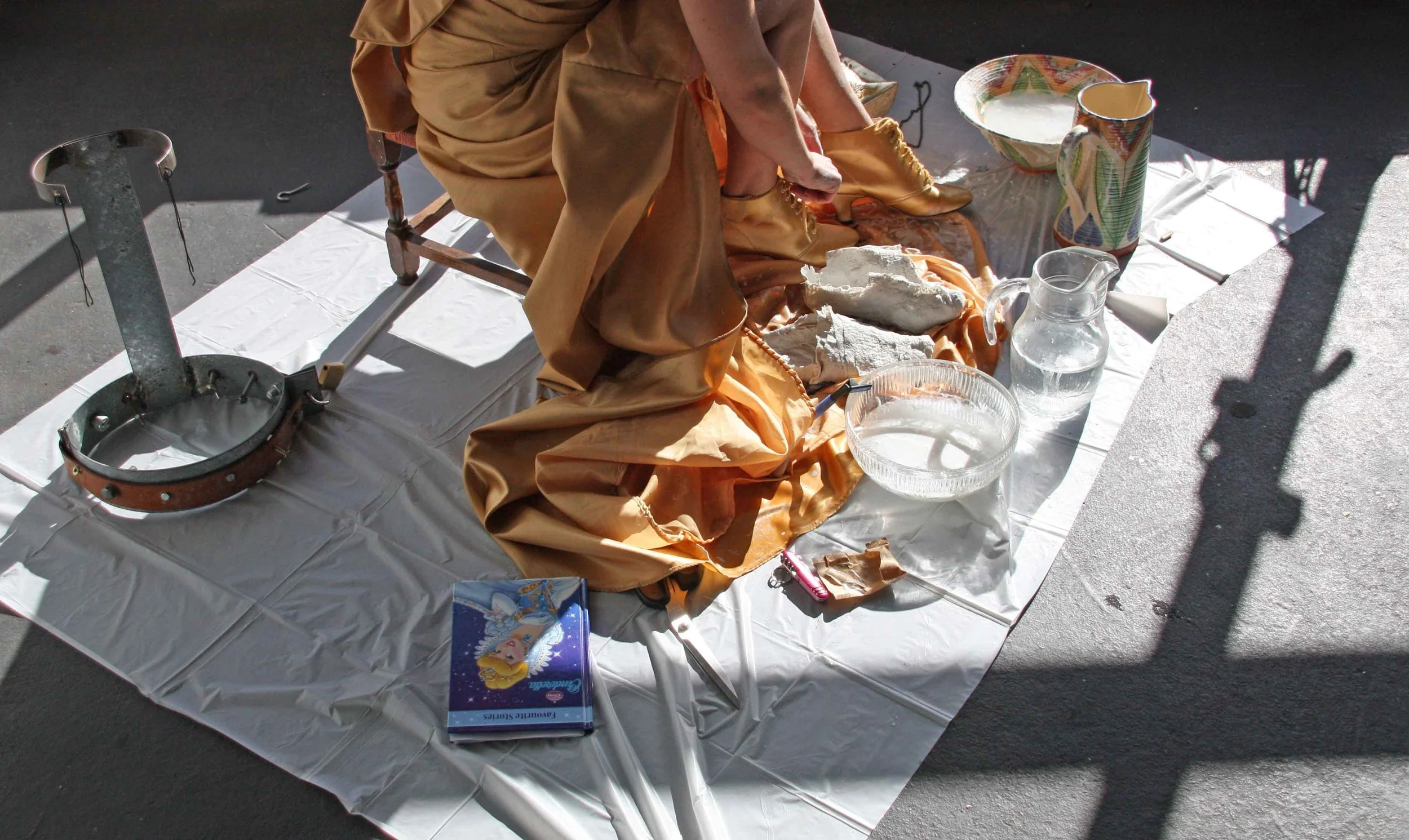 A person sitting on a small chair, is putting on a gold-colored boot. The person is seated on a white sheet spread on the ground, with various objects around, including a tambourine, a decorated ceramic pitcher, a glass pitcher of water, a small bowl