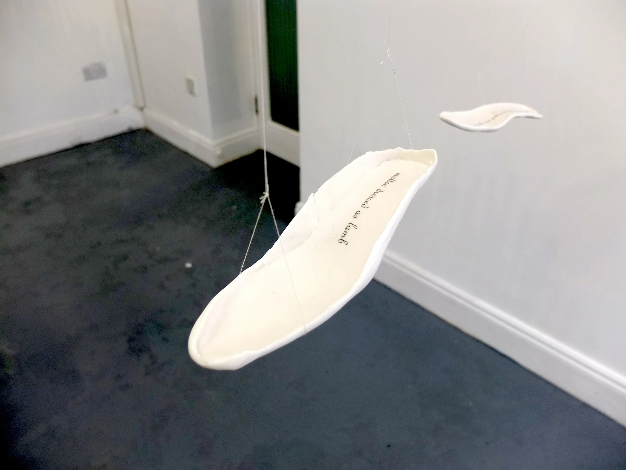Close-up of a white footprint made of ceramic hanging from thin strings against a white wall and black floor.