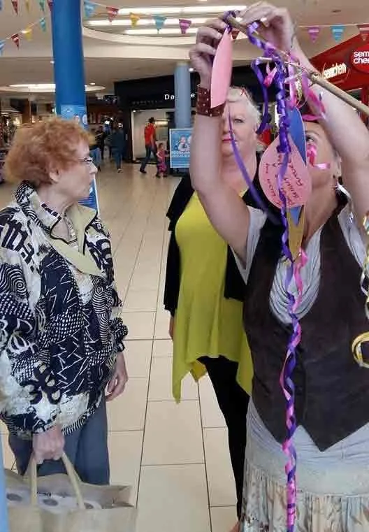Three women in a shopping mall, one is holding colorful ribbon decorations up in the air, while the other two are looking at her. The mall has festive banners and shops in the background.