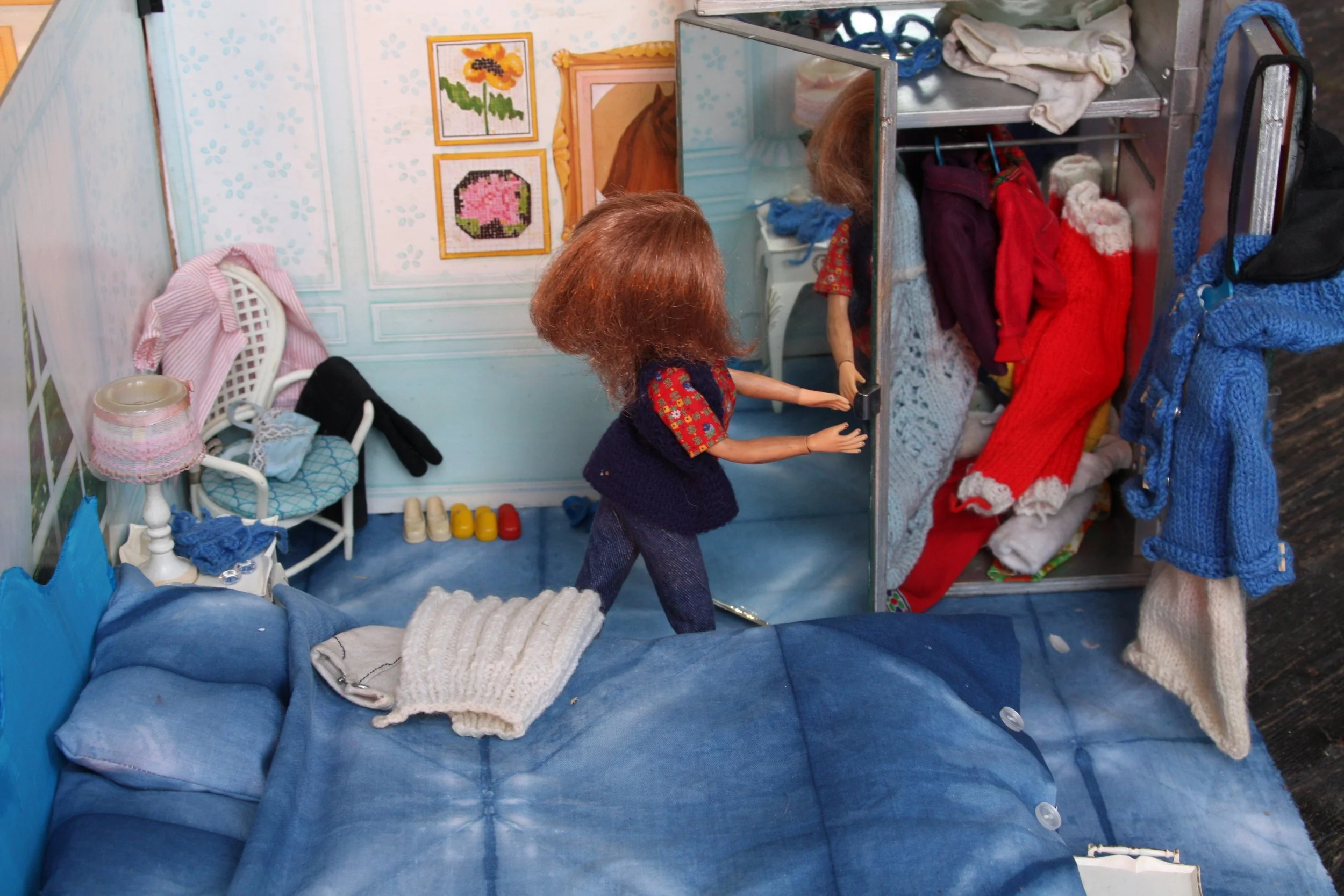 A dollhouse bedroom scene with a bed, a chair, a small table with a lamp, a wardrobe filled with clothes, and a mirror reflecting the room. The room has light blue walls with floral patterns and framed artwork.