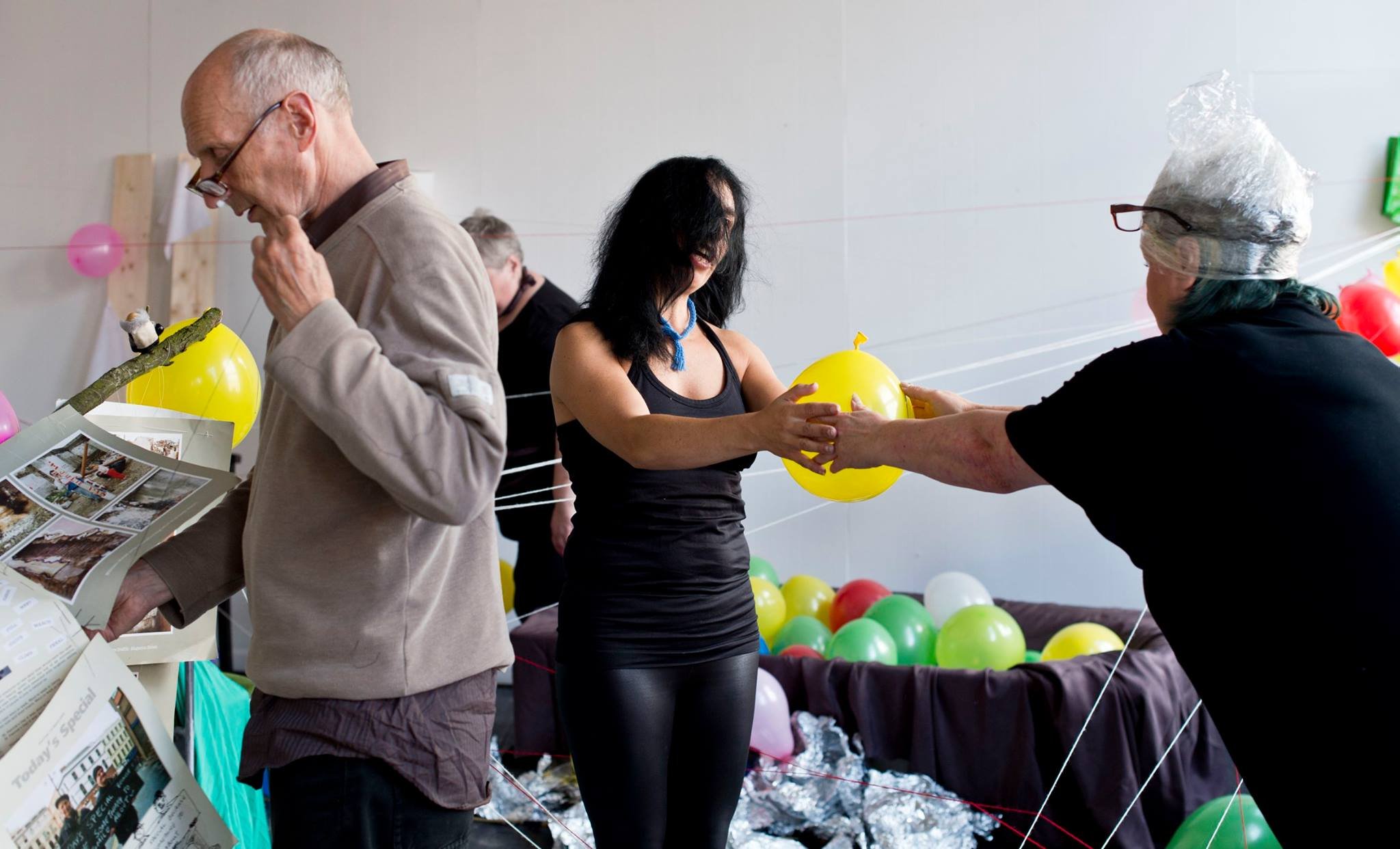 People at an event with colorful balloons. An elderly man is looking at a brochure, a woman with black hair and a necklace is holding a yellow balloon, and another woman wearing glasses and a head covering is taking the balloon from her. Other people