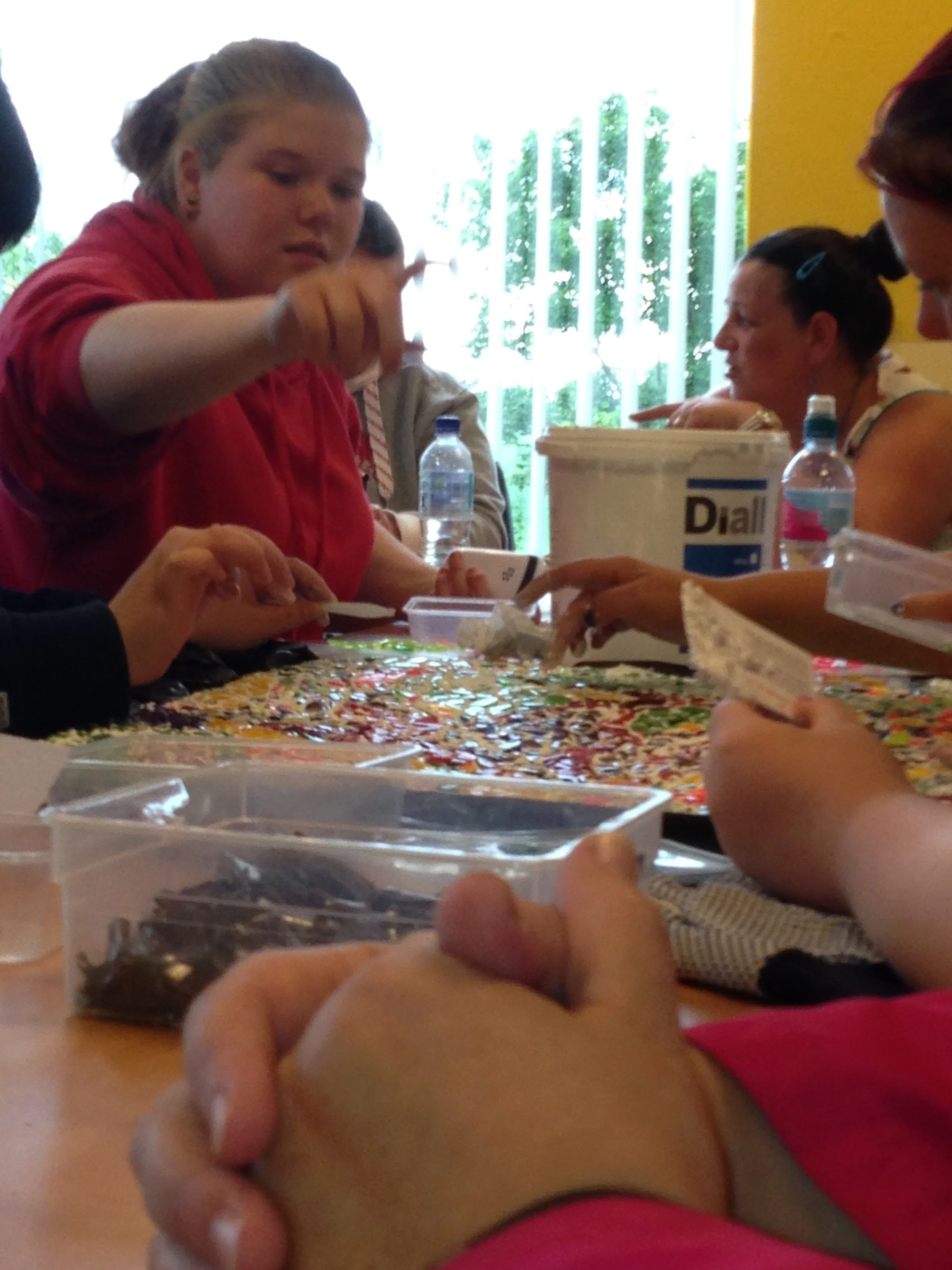 Several people sitting around a table engaged in an activity, with some holding small objects and others reaching into small containers, in a brightly lit room with large windows and green foliage outside.
