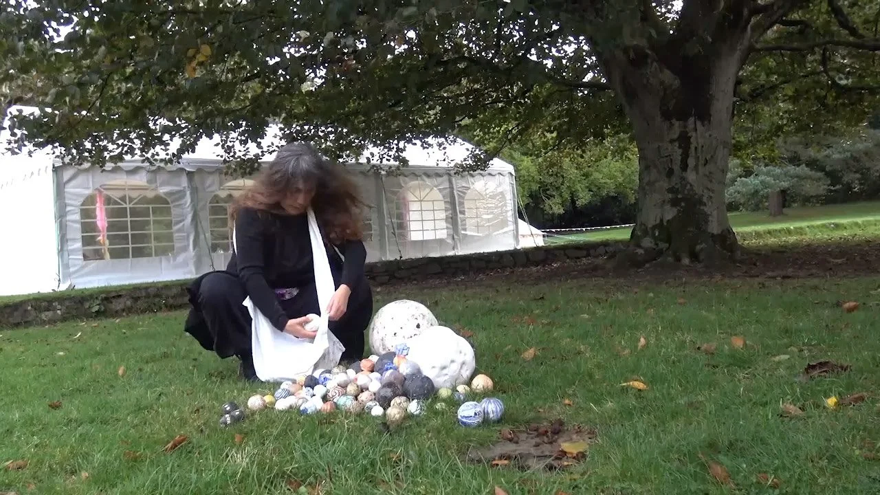 A woman with long dark hair, dressed in black, crouches down on a grassy area near a large tree, looking at various spherical objects and two large white ceramic sculptures on the ground.