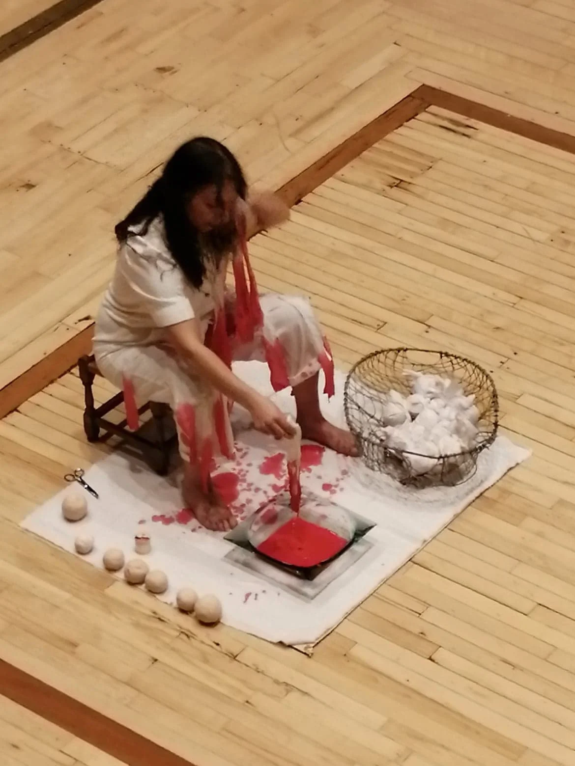 A person dressed in white is squatting on a small chair, pouring red liquid over their legs and feet, with red liquid spilling onto a white cloth beneath them. There are eggs and a scissors on the cloth, and a wire basket filled with white balls, pos