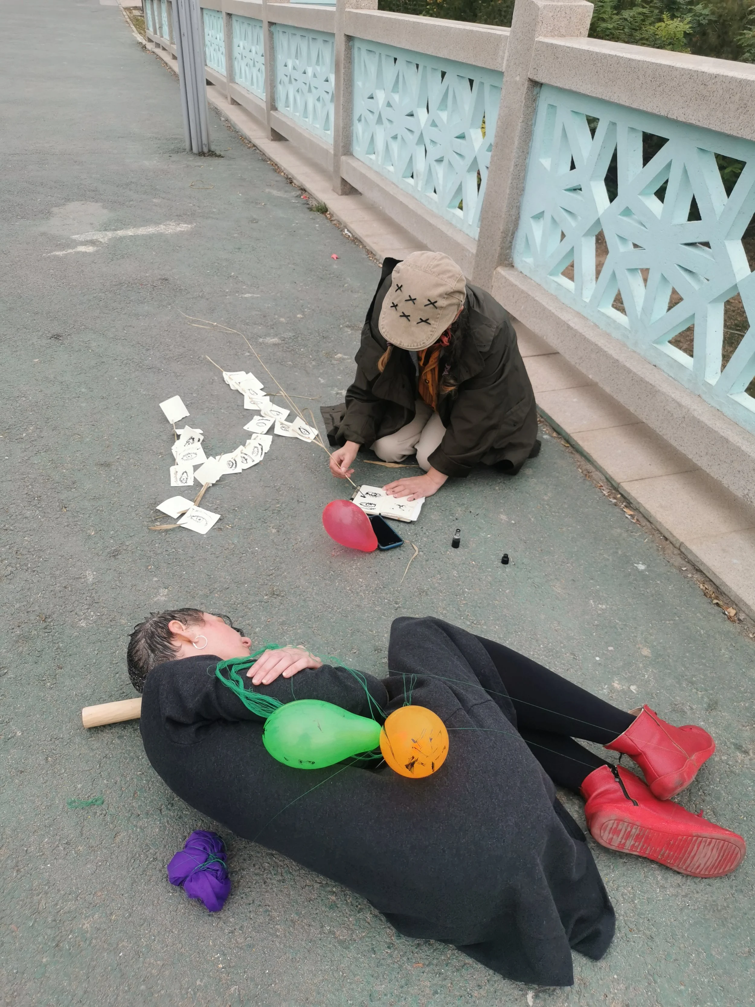 A woman lying on the ground with balloons around her, another person crouched nearby with face obscured, and a third person sitting with closed eyes wearing a hat and jacket, beside a fanciful blue and white fence.