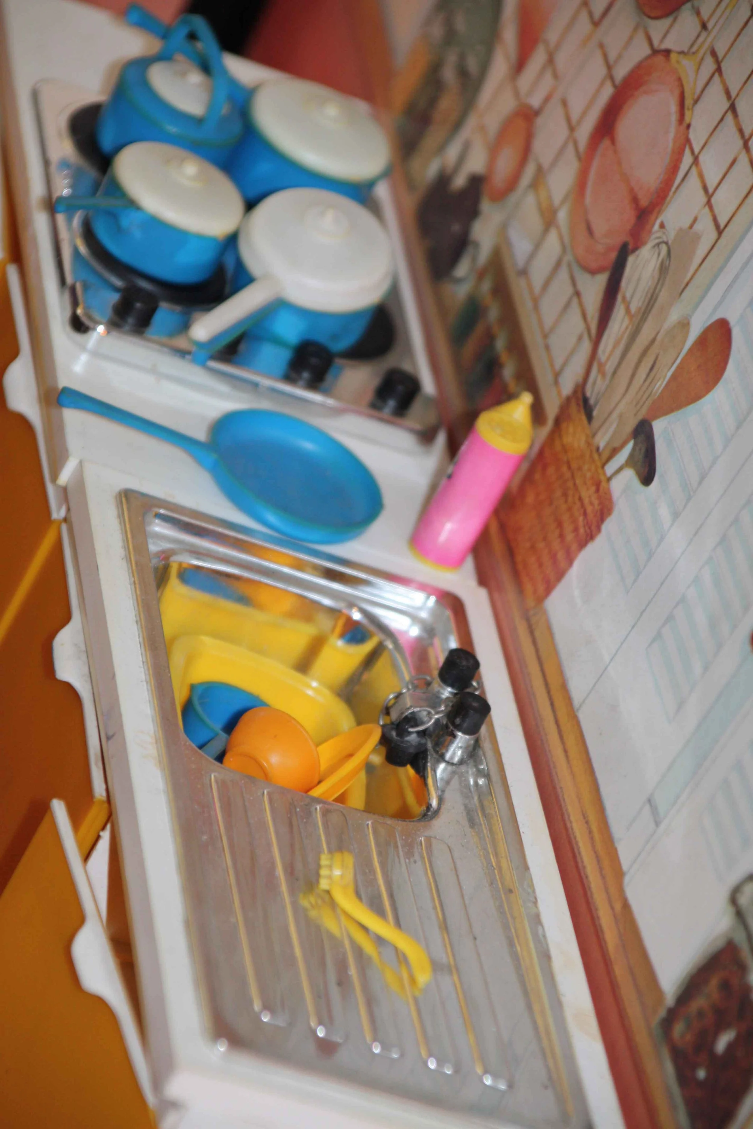 Miniature toy kitchen sink with yellow utensils, orange ball, blue ladle, and gray and black toy knobs, with a colorful kitchen-themed wallpaper in the background.