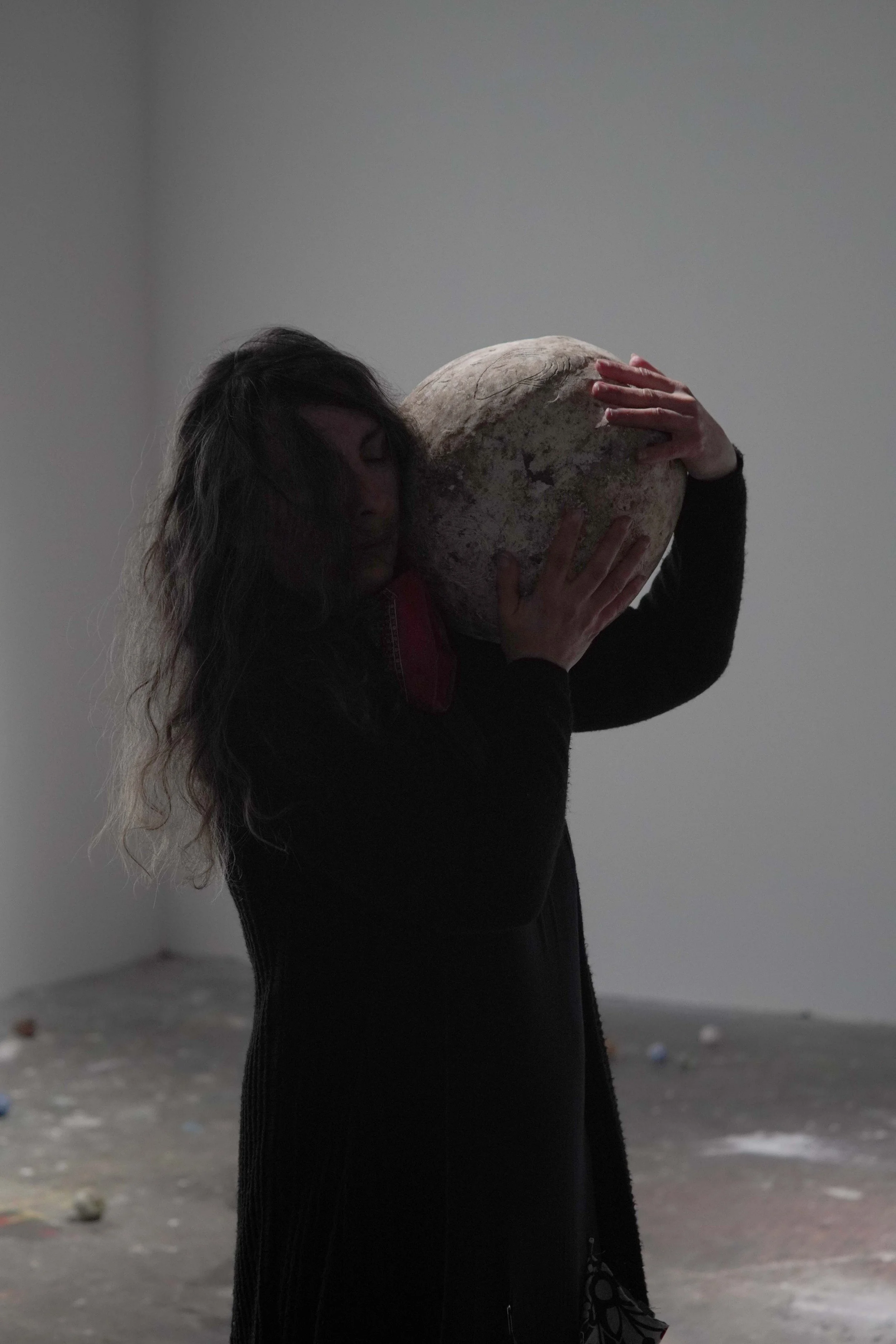 A woman with long dark hair, wearing a black outfit, is holding and embracing a large, round stone or object against her chest, with her face resting on it. The background is a plain, light-colored wall, and the ground appears to be a concrete or gra