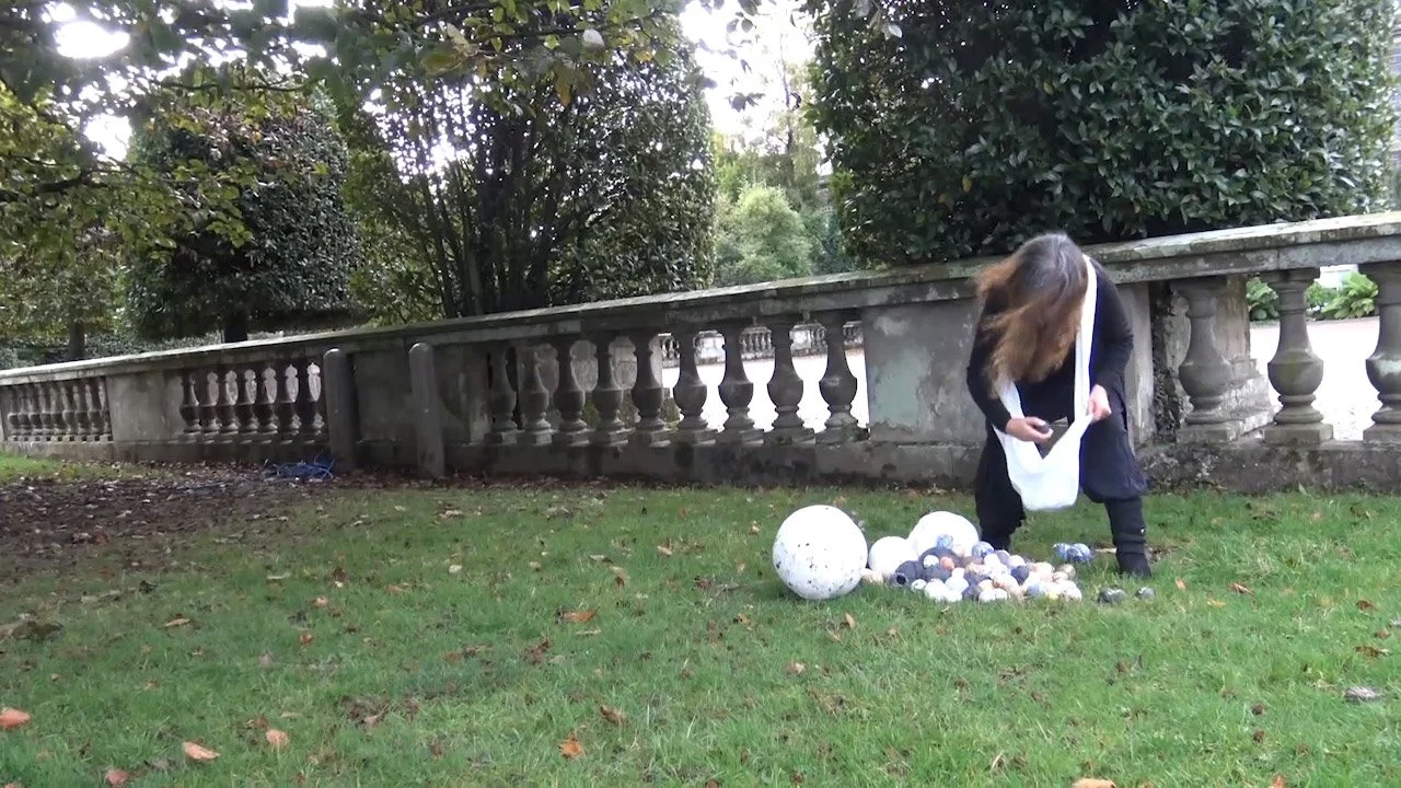 Person bending over a pile of small and large ceramic balls on a grassy area, with a stone fence and trees in the background.