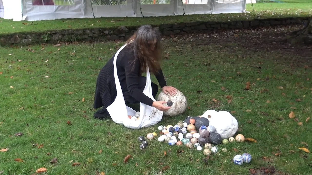 A woman kneeling on the grass outdoors, surrounded by various painted and unpainted ceramic balls, some resembling eggs, and a large white decorative object, with a large speckled ball and a smaller item in her hand.
