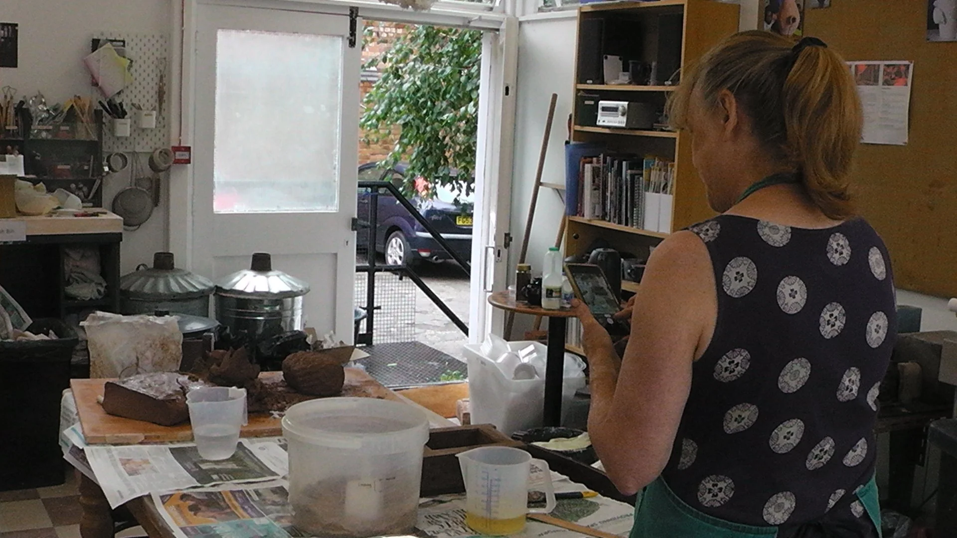 A woman in a sleeveless dress with a circular pattern looks at her phone inside a busy kitchen or workshop. The counter in front of her has various containers, newspapers, and equipment, with an open door revealing a parked car and greenery outside.