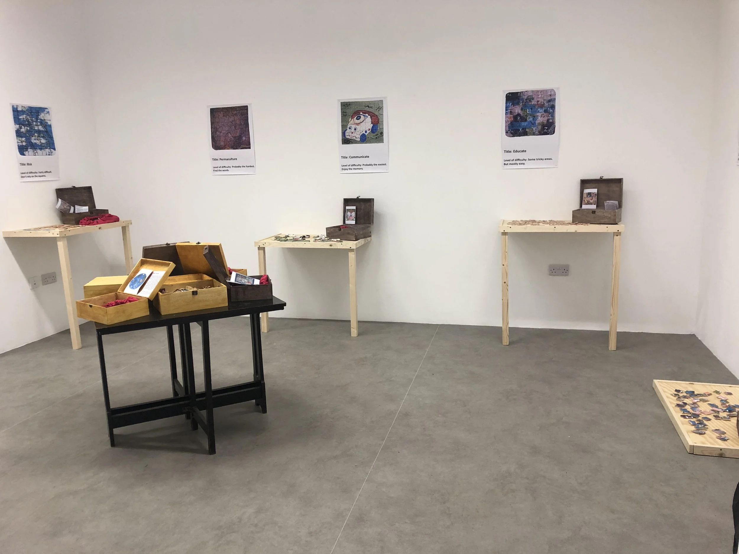 An art exhibition with puzzle-themed artwork and informational posters on the wall, and wooden display tables with puzzle pieces and boxes.