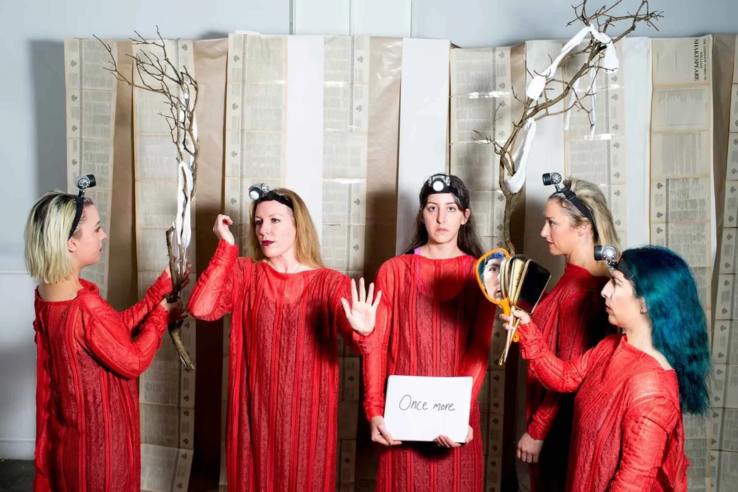Six women dressed in red, standing in front of a decorated folding screen, some holding branches with white cloth strips, one holding a sign that says 'Once more'.