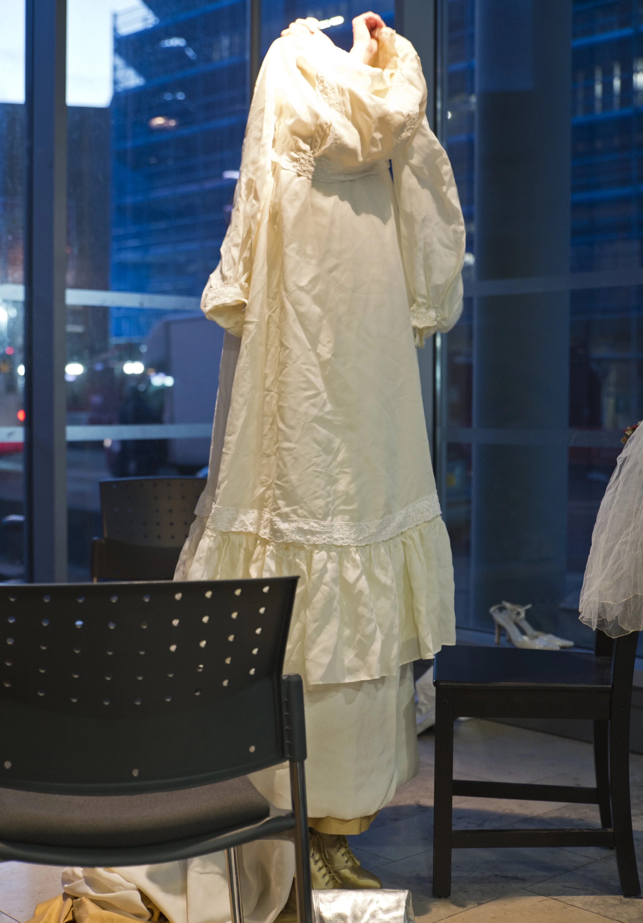 A vintage wedding dress on a mannequin inside a glass storefront with city buildings in the background.