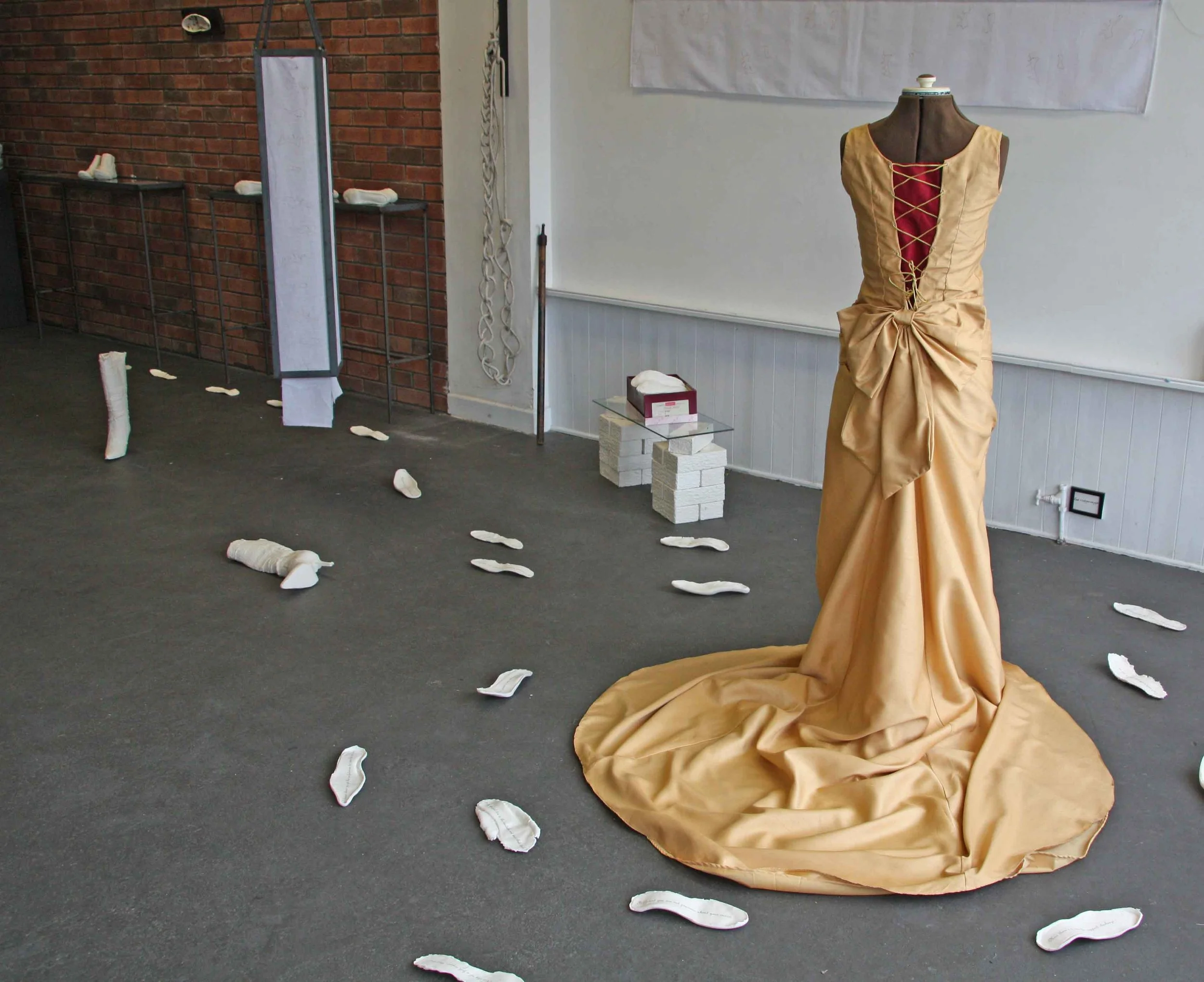 A dress form with a gold satin dress in a room with a brick wall and whiteboard. Scattered fabric pieces on the floor.