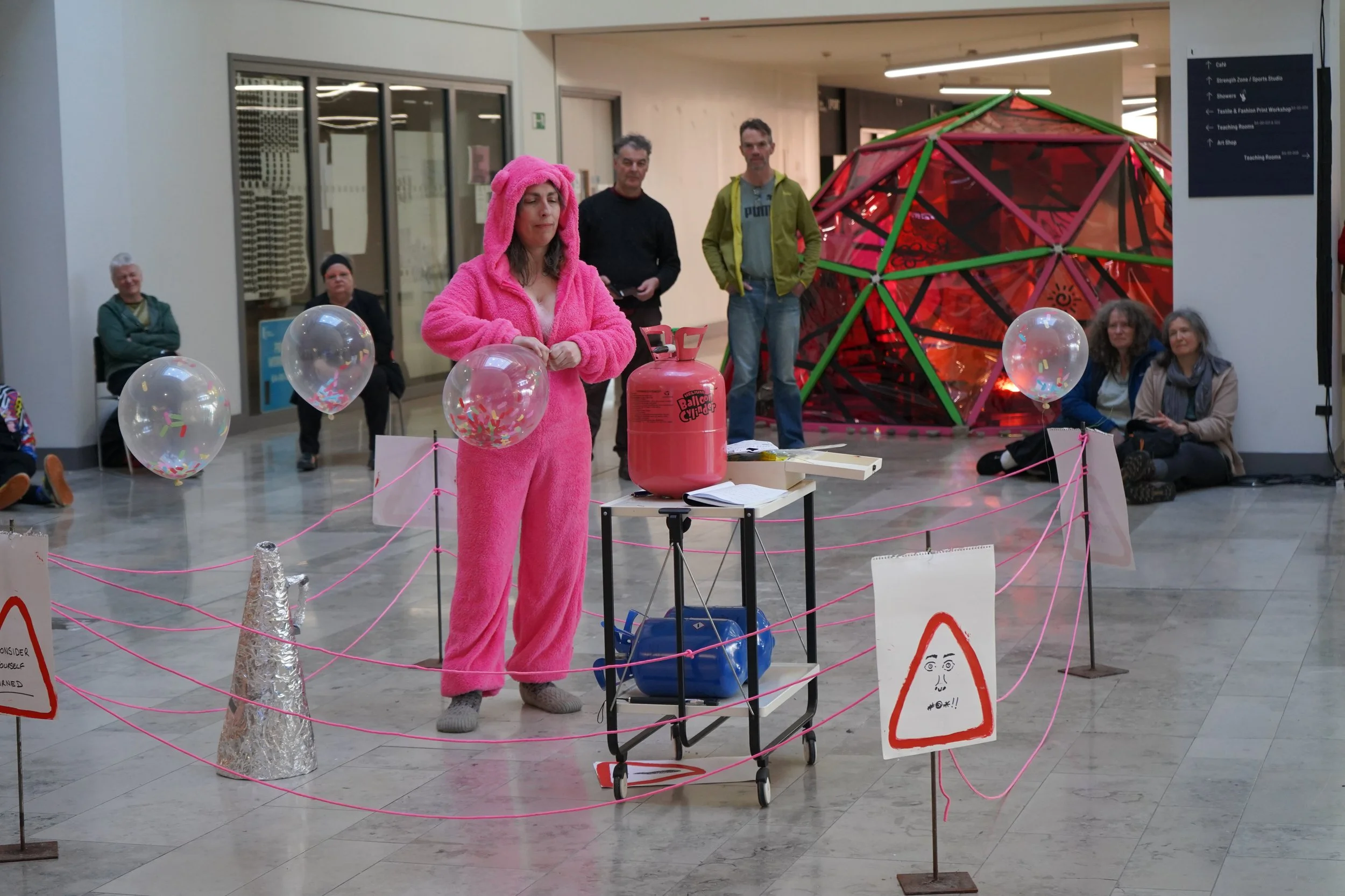 A woman dressed in a pink furry onesie with a hood, resembling a rabbit, standing behind a table with a pink helium tank and balloons, in a public indoor space with other seated people and a colorful geodesic dome in the background.
