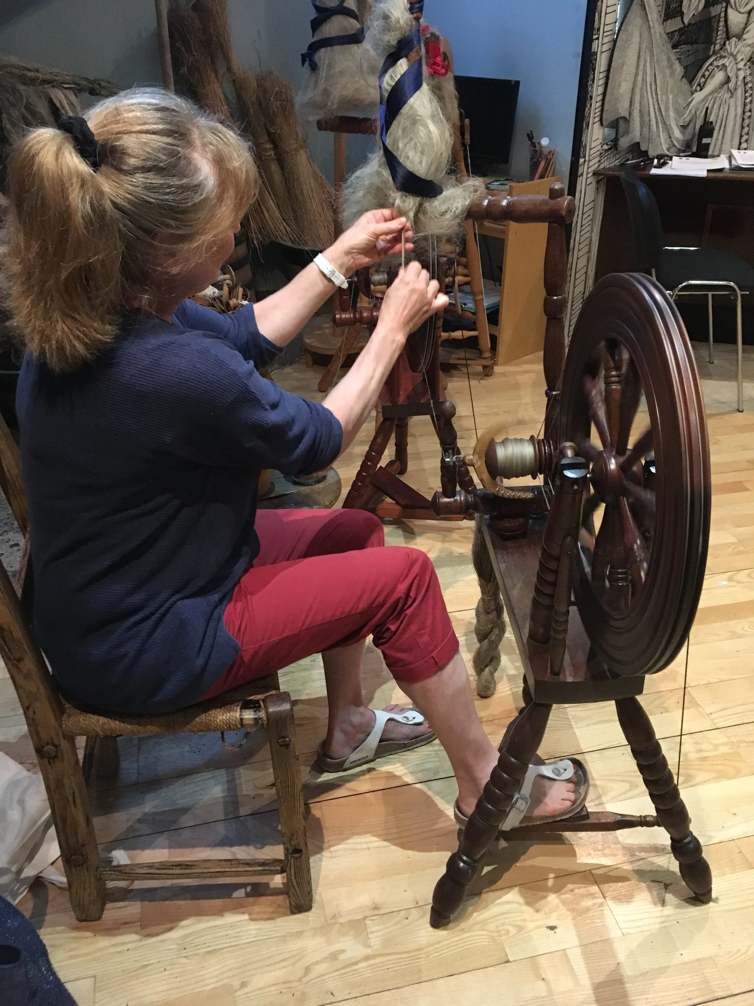A woman with blonde hair tied back in a ponytail, wearing a navy blue top, red pants, and white sandals, is seated on a wooden chair. She is working on a large, woven spinning wheel, which is used to spin fiber into yarn. The room appears to be a cra