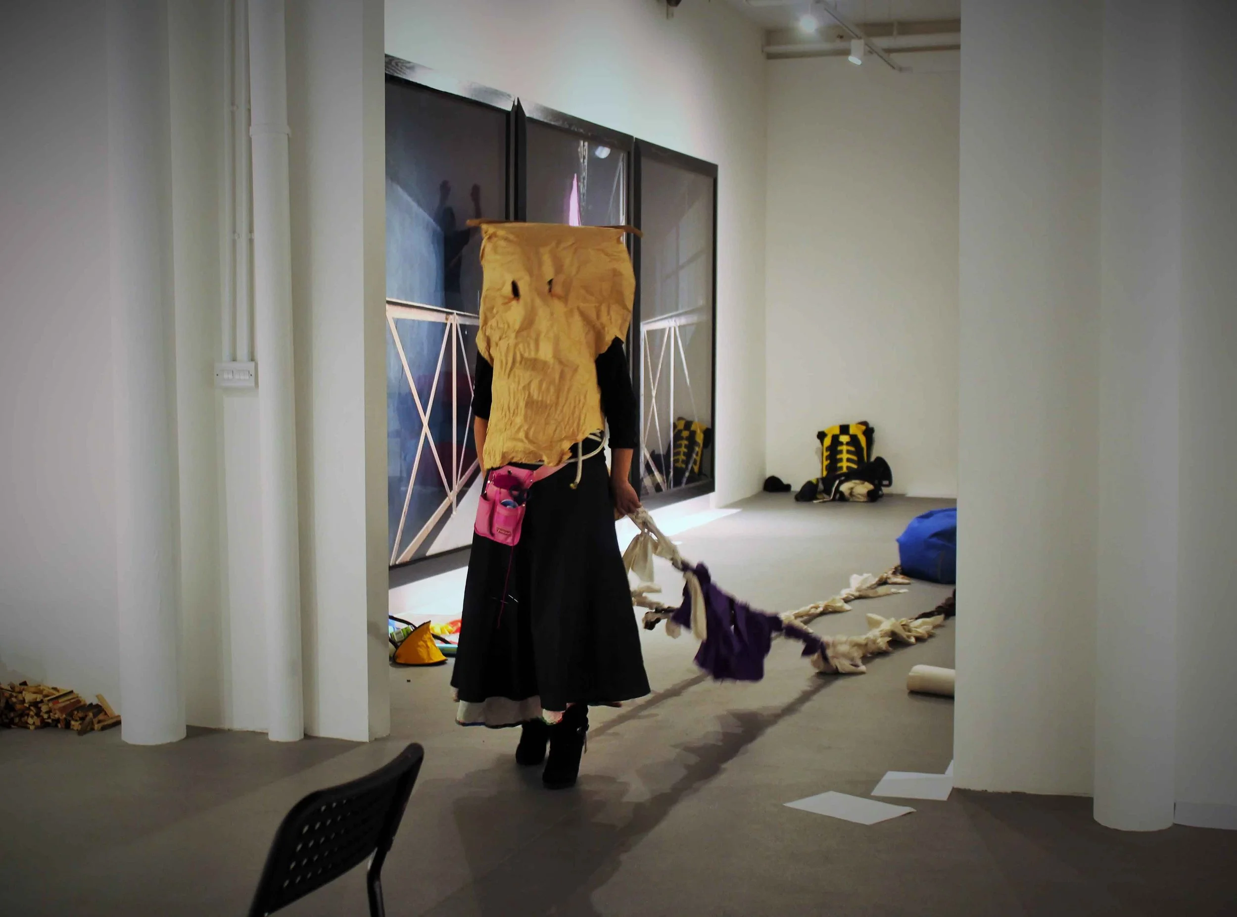 Person dressed in costume with a paper bag on their head, walking with a dog inside a modern indoor space with artwork on the wall and chairs on the floor.