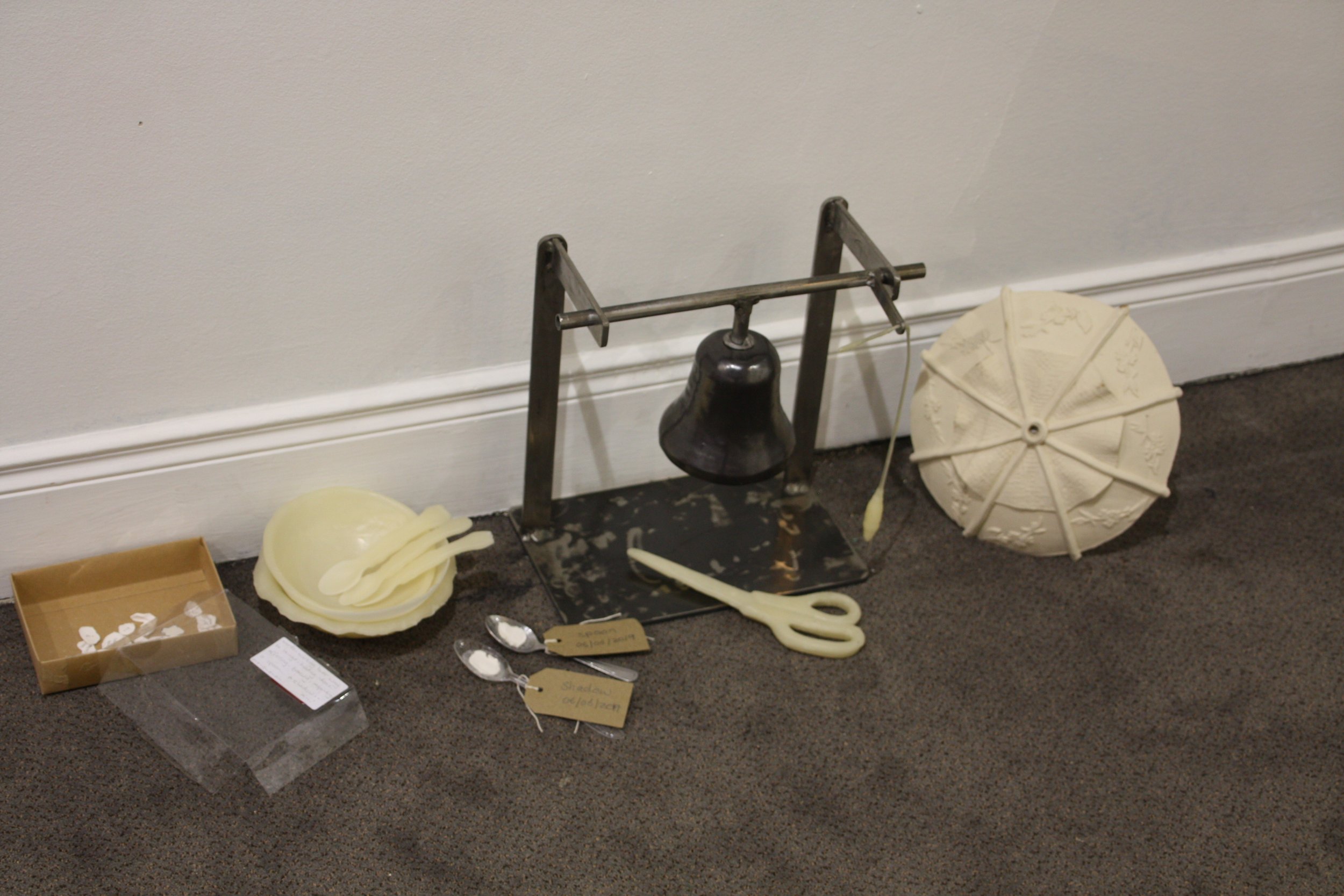 A metal mold set-up for making items in a craft or art project, with a large wheel, scissors, and various crafting materials on the floor, against a plain wall.